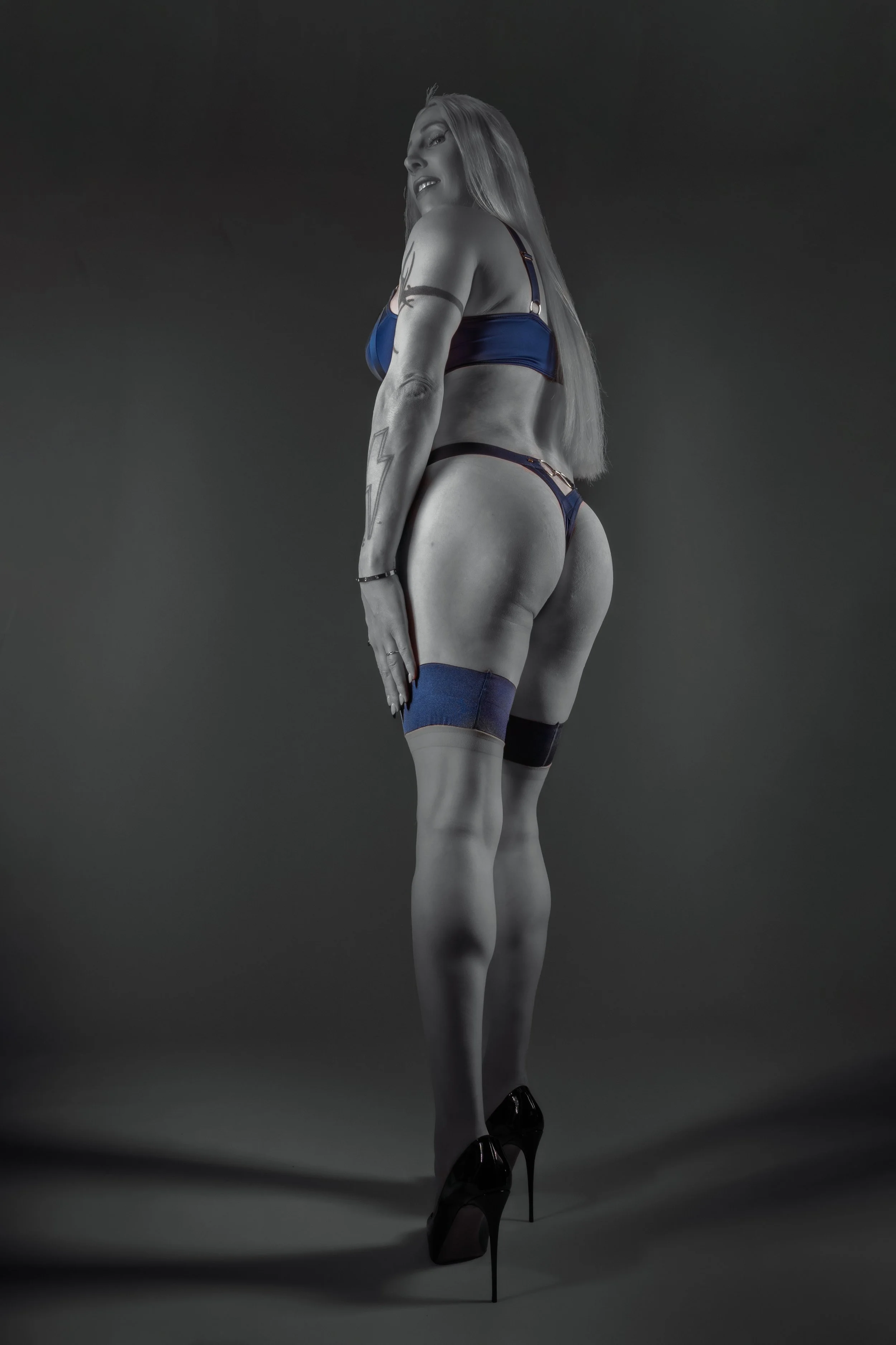 A woman in high heels and lingerie, standing with her back turned, looking over her shoulder in a studio with a gray background.