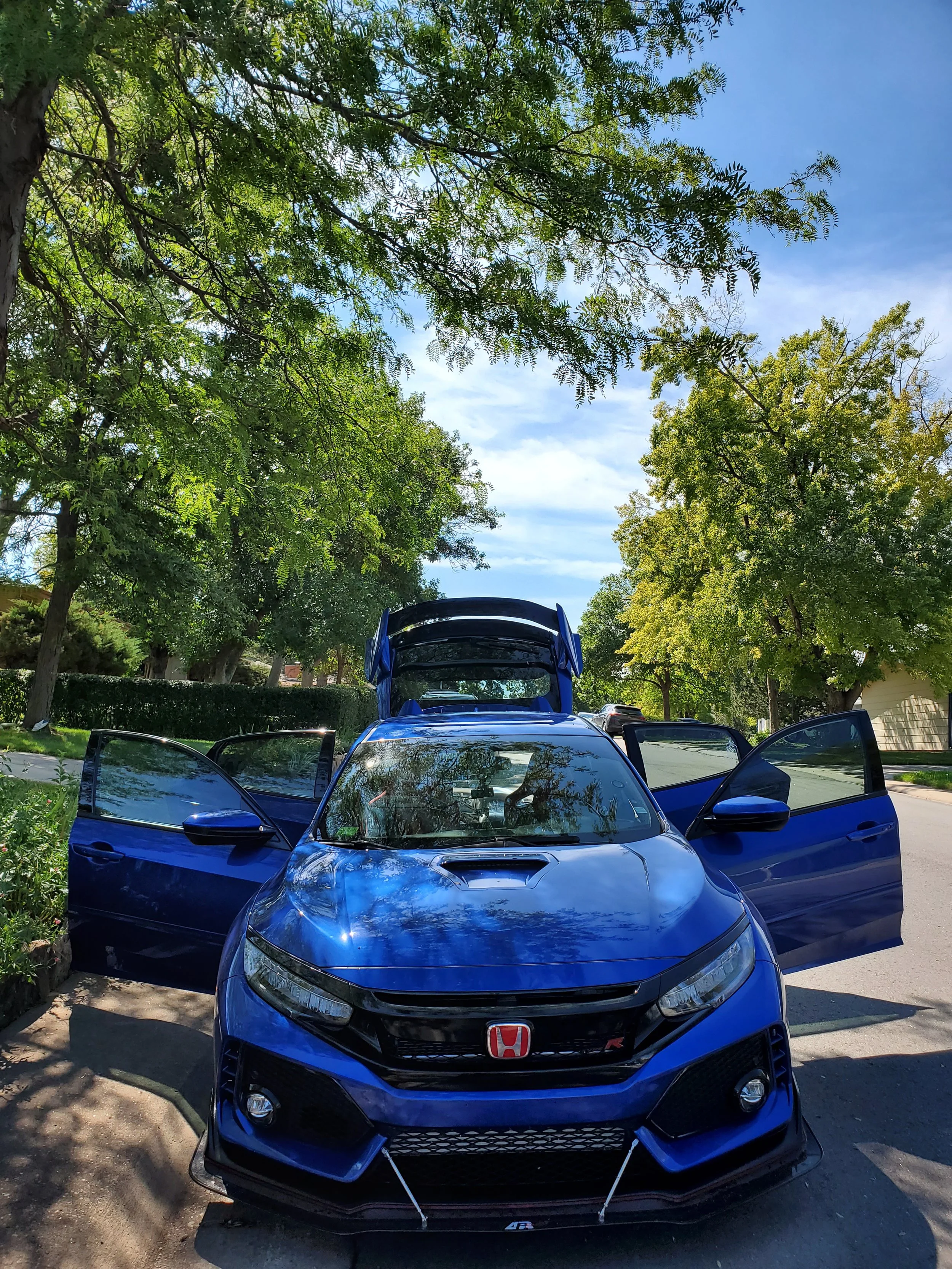 A blue Honda Civic Type R with doors open and a rear hatch lifted, parked on a street with trees and a blue sky in the background.