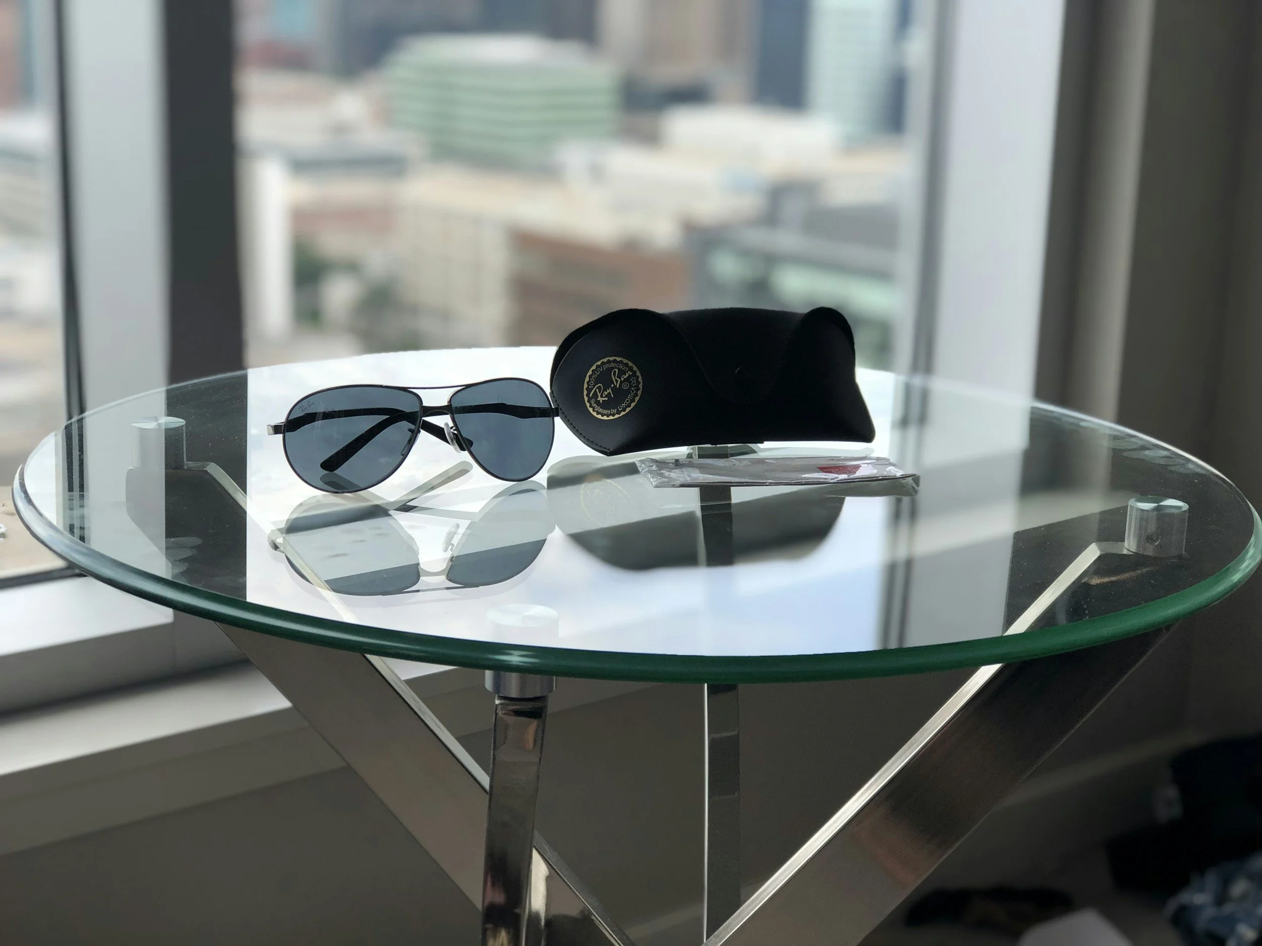 A pair of Ray-Ban sunglasses and a black Ray-Ban case on a glass table in front of a window with a cityscape view.