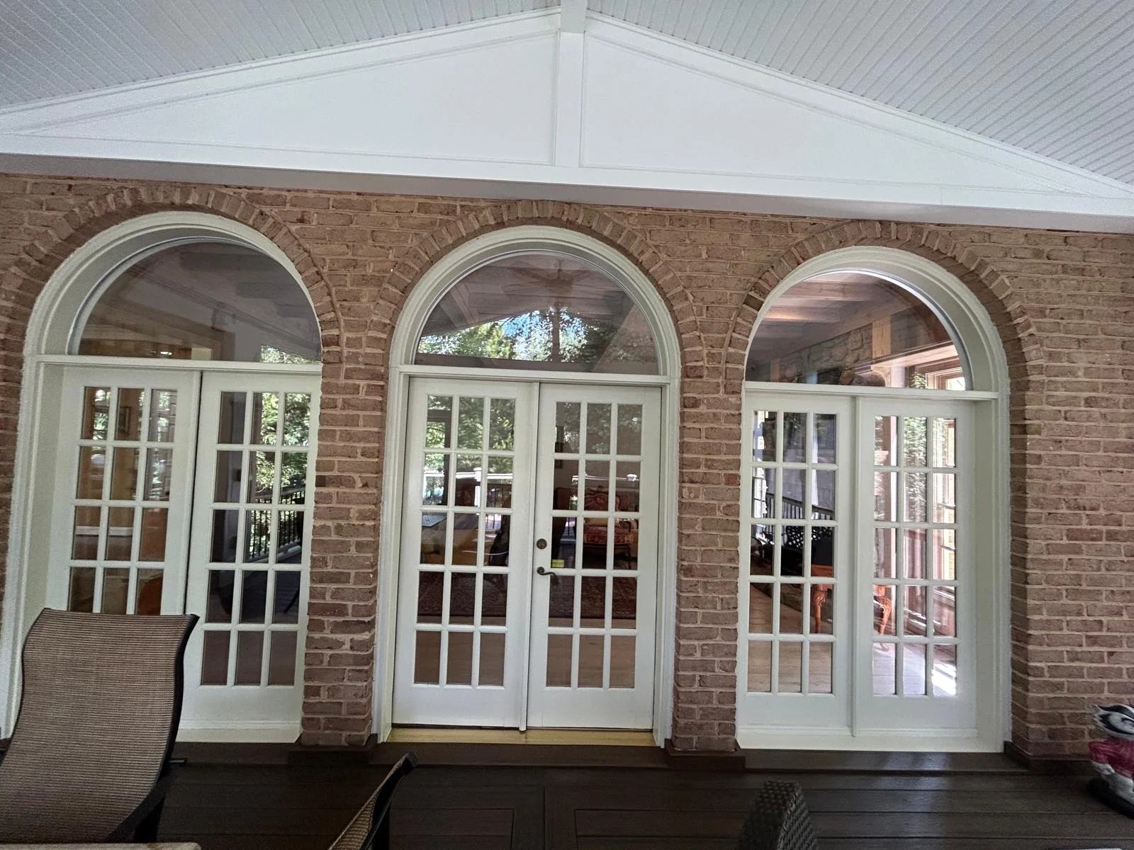 Three arched glass-paneled double doors with white frames set in a brick wall, leading into a room with wooden flooring and visible furniture, viewed from a porch with dark wood flooring.