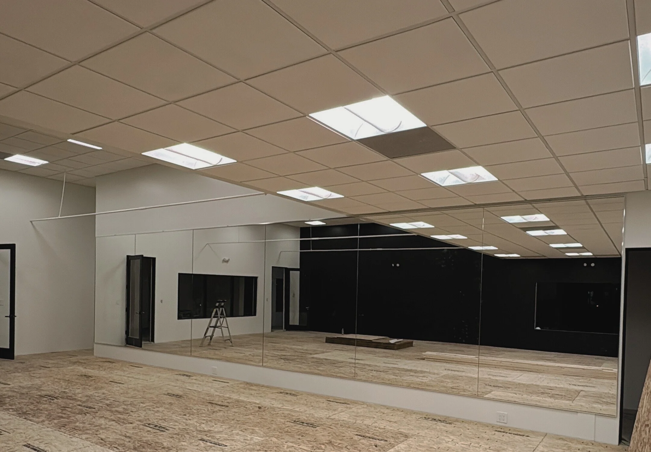 An interior room with a large mirror wall, exposed ceiling tiles with fluorescent lights, a ladder, and a black accent wall. The floor is unfinished with construction materials.