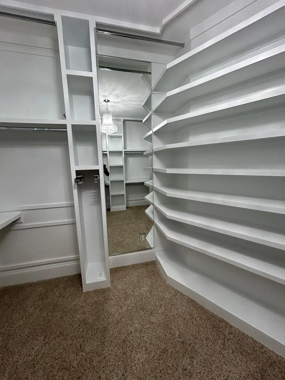Empty walk-in closet with white shelving units, a full-length mirror, and a chandelier reflected in the mirror. The closet has beige carpet flooring.