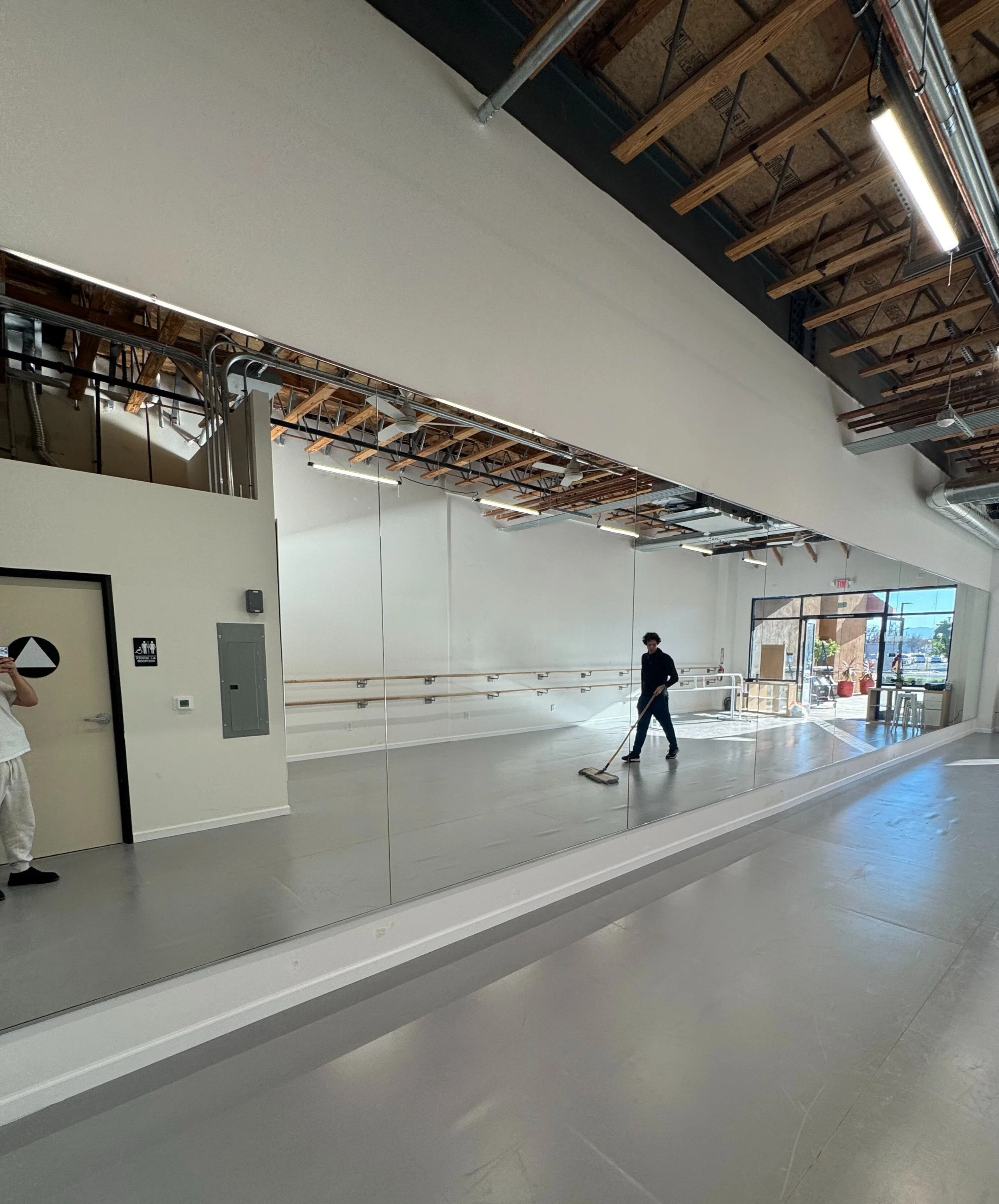 A person sweeping the floor inside a spacious, empty dance studio with mirrored wall, ballet barres, and large front window with sunlight streaming in.