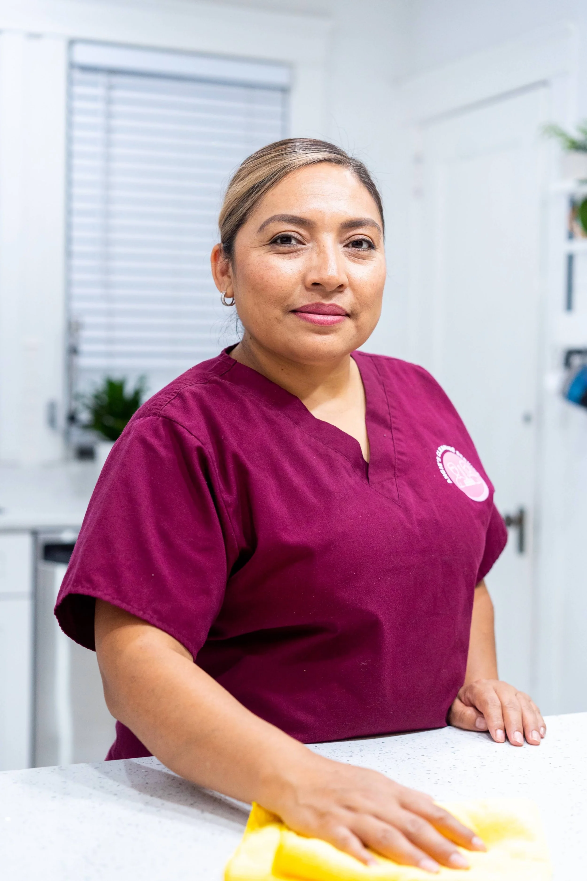 A woman wearing a maroon scrub top standing at a white countertop with a yellow cloth in her hand, in a bright white room, possibly a kitchen or medical office.