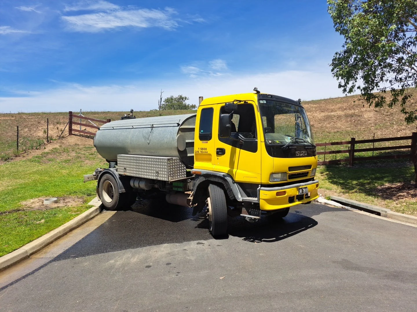 A yellow Isuzu water truck with a silver tank is parked on a paved area near a grassy lawn, with a wooden fence and a hill in the background under a partly cloudy blue sky.