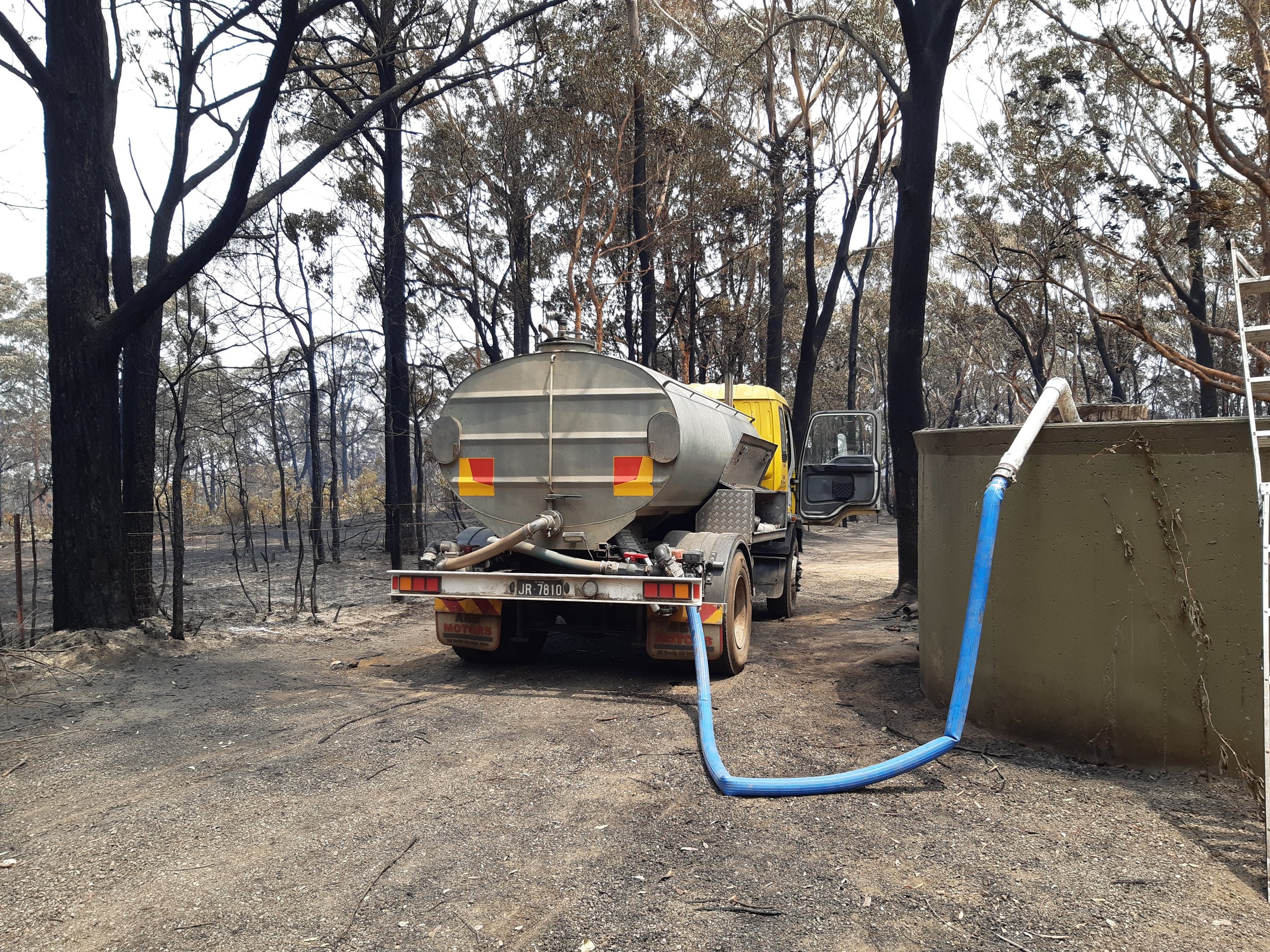 A water tanker truck parked on a dirt area surrounded by burnt trees, with a hose connected to a large tank on the right side.