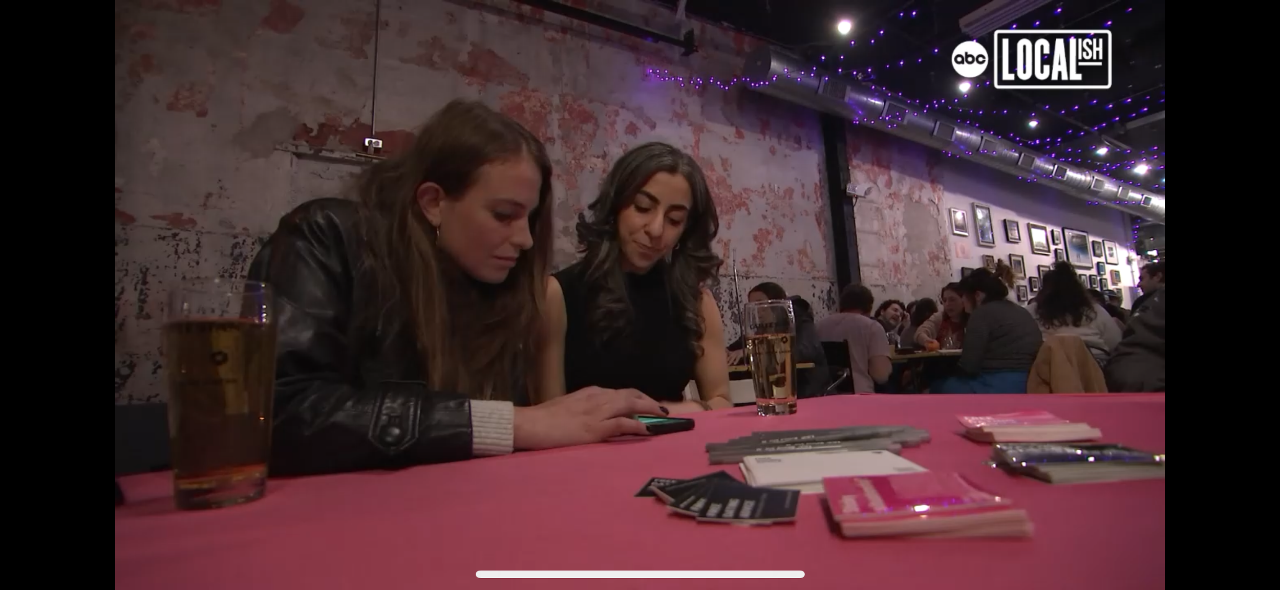 Two women sitting at a table in a crowded bar or restaurant, looking at a phone. There are drinks and cards on the pink tablecloth. The background has an exposed brick wall with framed pictures and purple string lights.