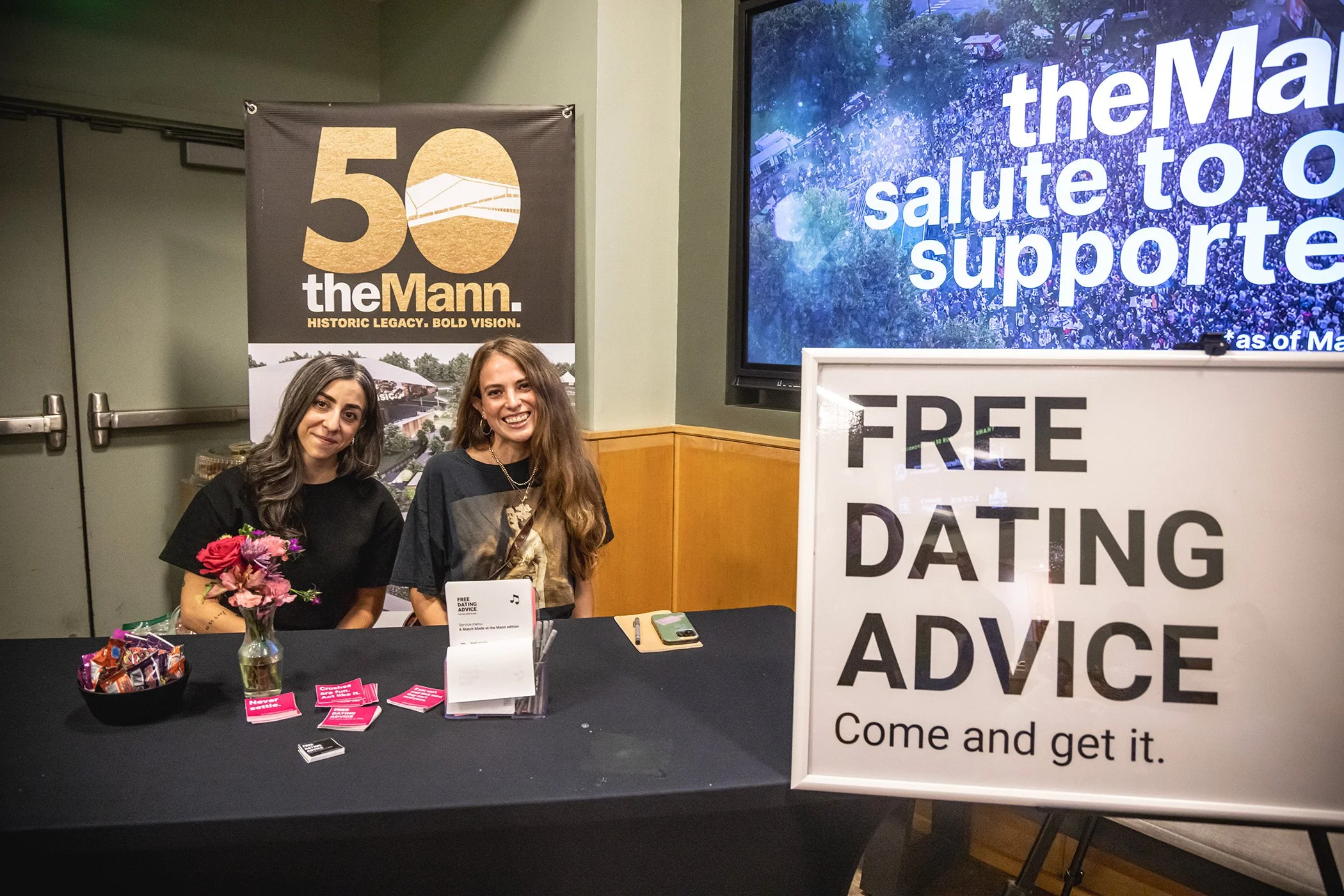 Two women sitting behind a table with a sign offering free dating advice at an event, with a large screen and banners in the background.