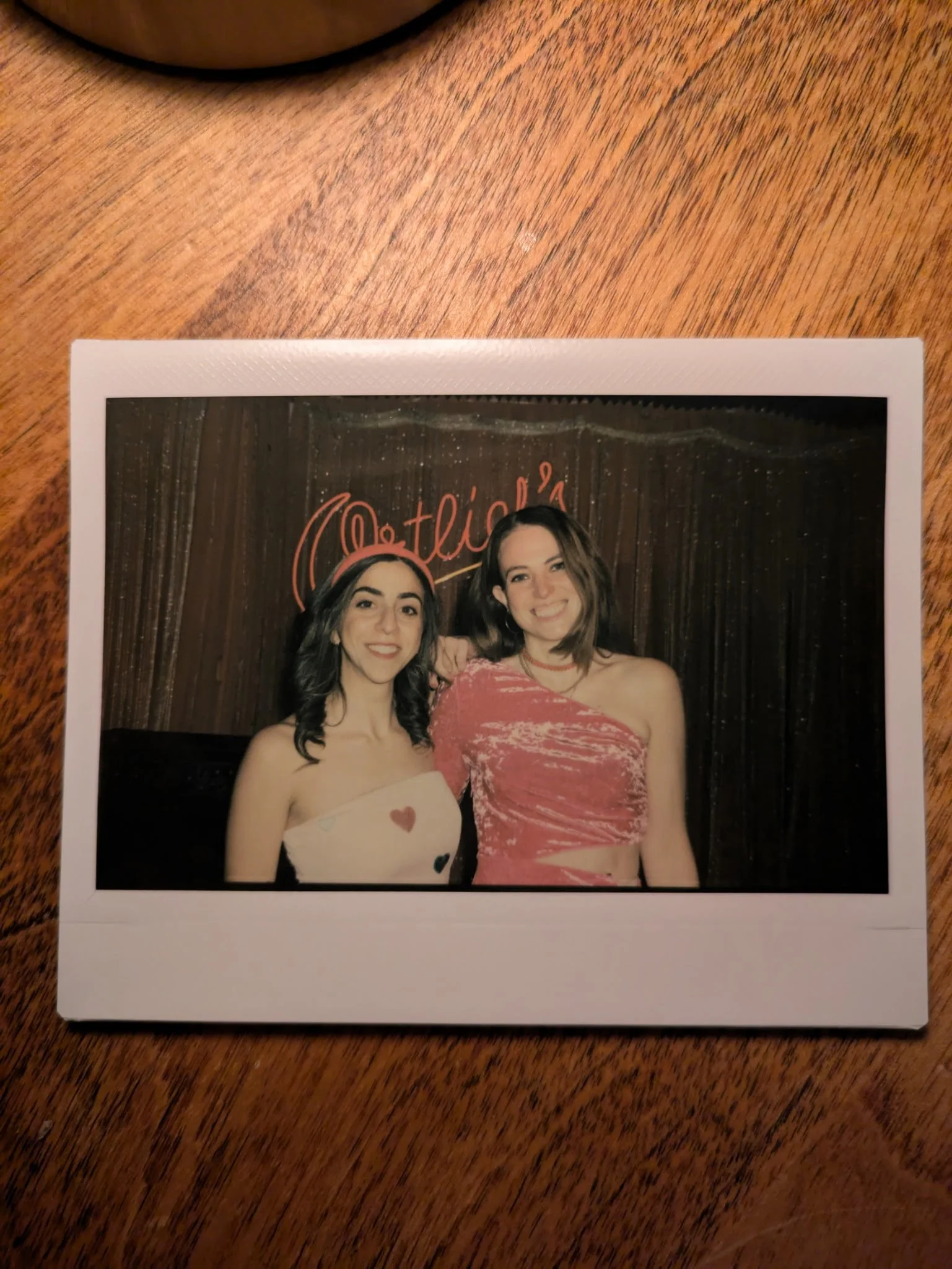 Two women smiling and posing in front of a dark curtain with a red neon sign, one in a strapless top with hearts, the other in a one-shoulder pink dress, inside a photo booth.