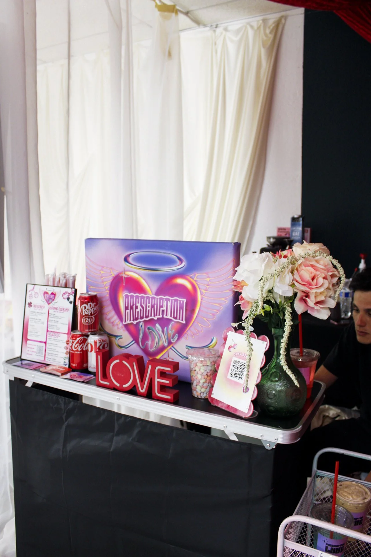 A table decorated with a large pink and purple heart sign that says 'Resignation Love', a green vase with pink and white flowers, love-themed decorations, drinks, and a QR code. Curtains in the background.