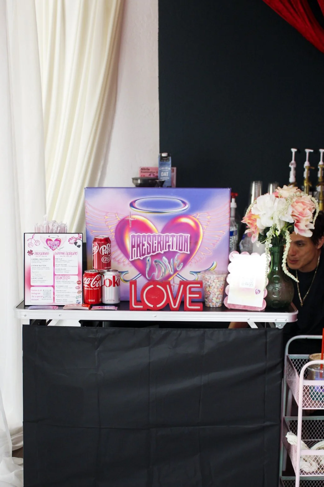A decorated table for a Valentine's Day-themed event with pink and red accents, featuring a sign that says "Preservation Love" and "Love," pink flowers, Coca-Cola cans, marshmallows, and a menu, with a person partially visible on the right.