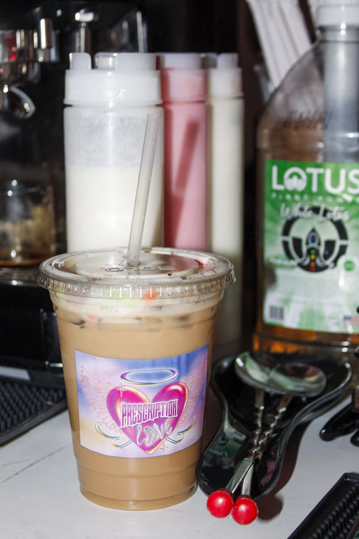 A plastic cup with a lid and straw containing iced coffee. The cup has a sticker that says 'PRESCRIPTION LOVE' with a heart and angel wings. In the background, there are condiment bottles, including sugar and flavored syrups, and a pair of black scis