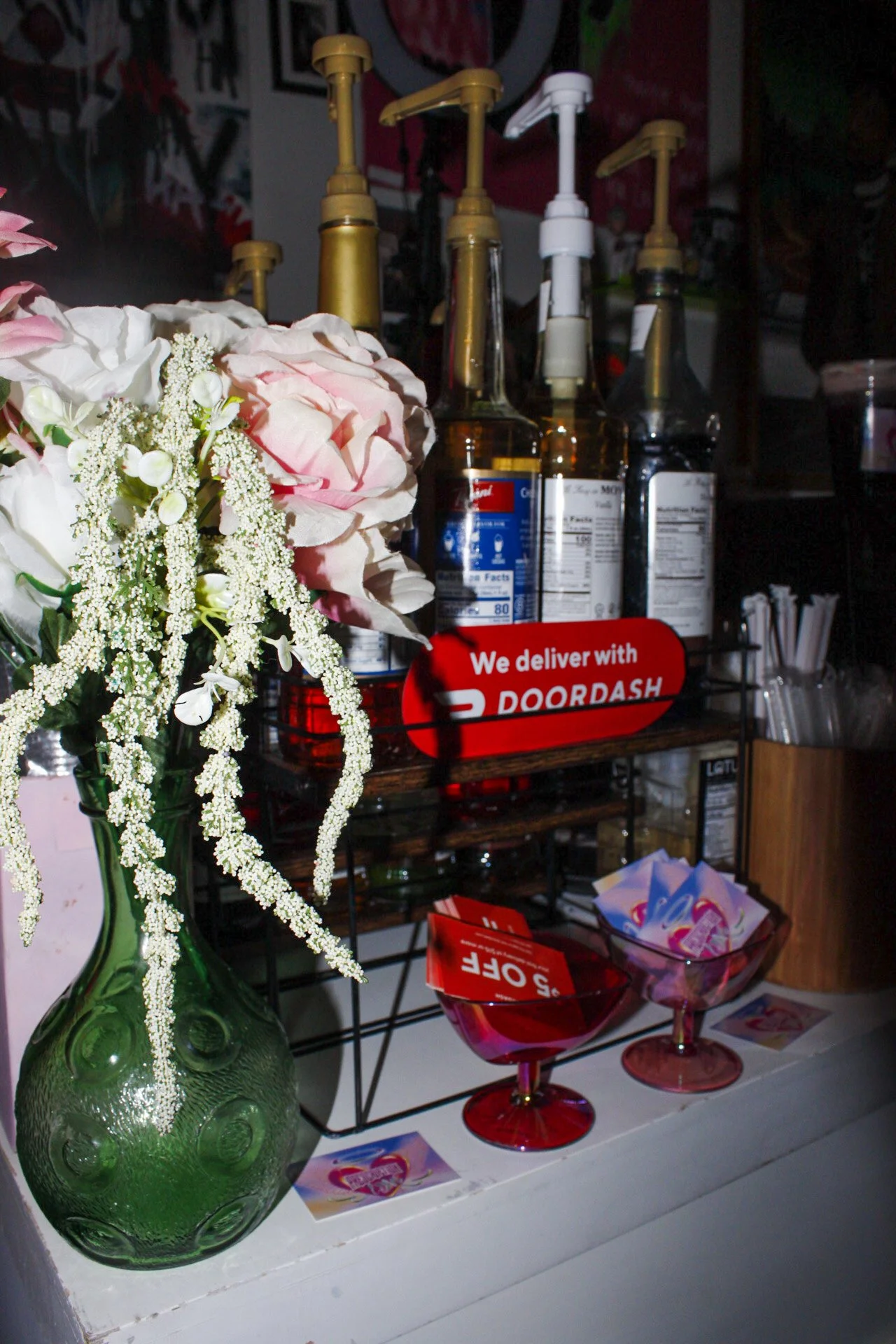 A green vase with pink and white flowers, several bottles of syrup or flavoring, a sign that says 'We deliver with DOORDASH', and pink containers with red and white sale tags on a white surface.