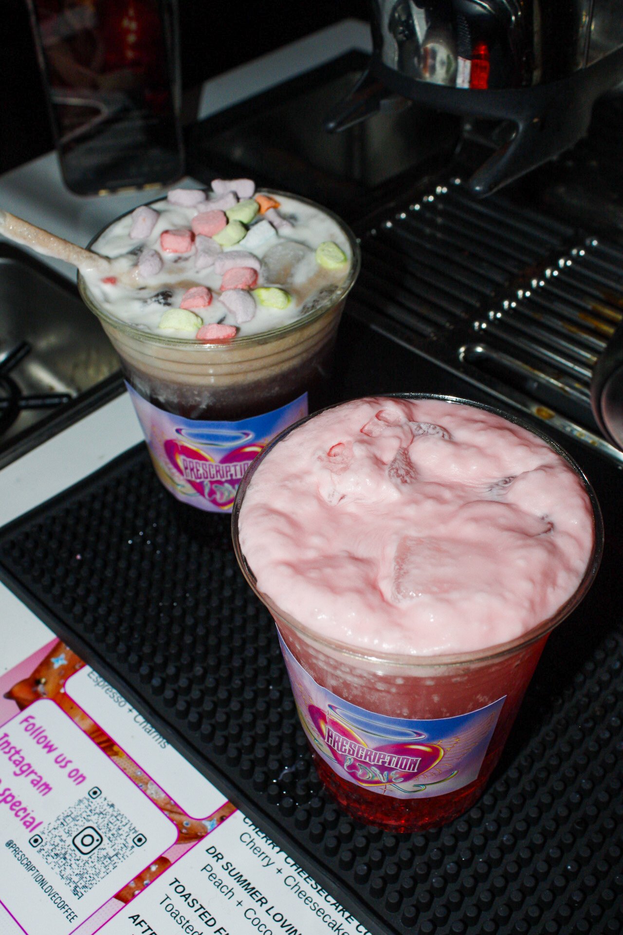 Two glasses of pink and brown frozen drinks topped with marshmallows on a black rubber mat on a kitchen counter.