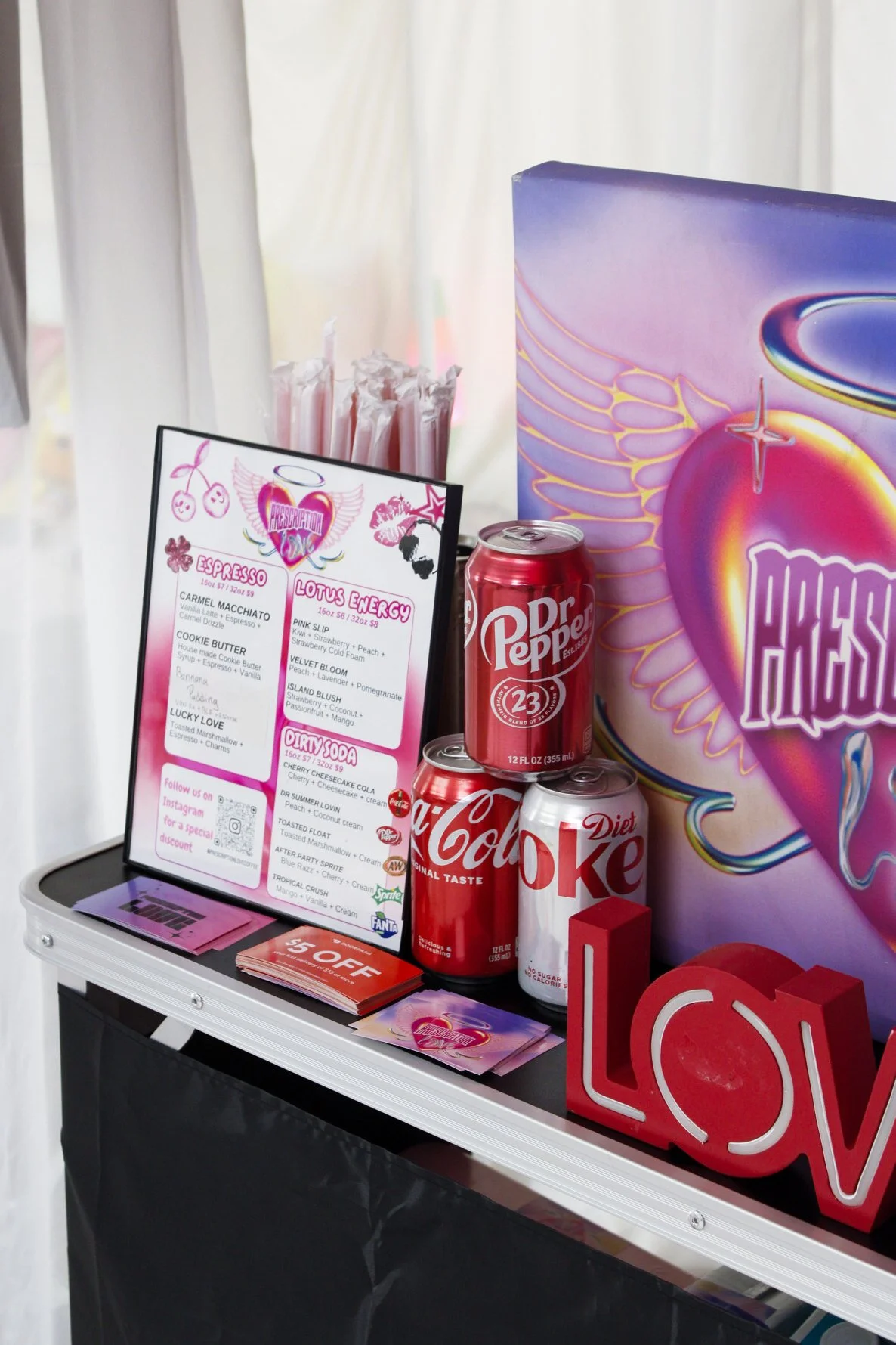 A display table with canned soda, a menu, a large decorative heart with wings labeled 'Hugs', and love-themed decorations.