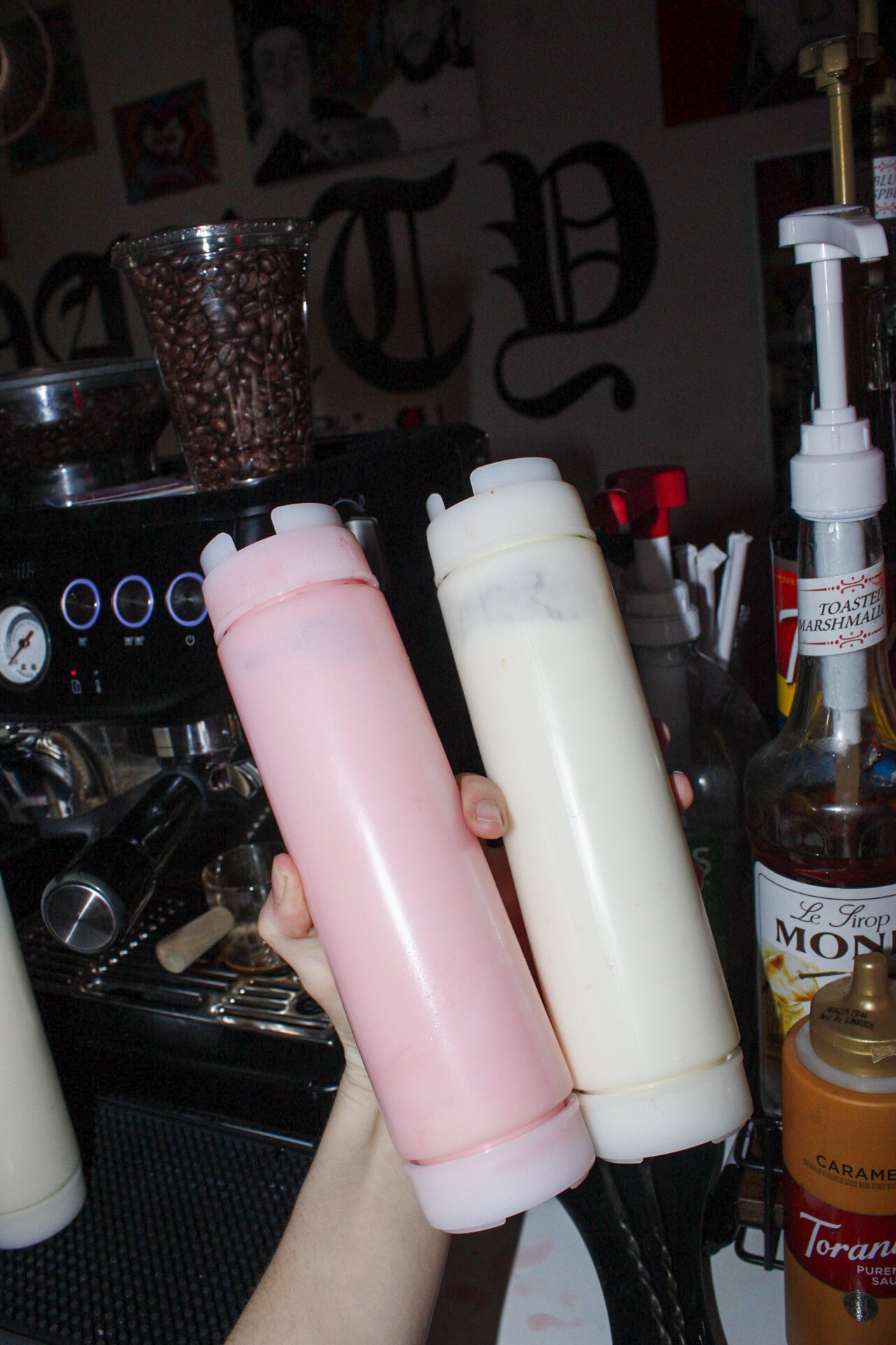 Person holding two condiment bottles, one pink and one white, in a coffee shop or cafe setting."