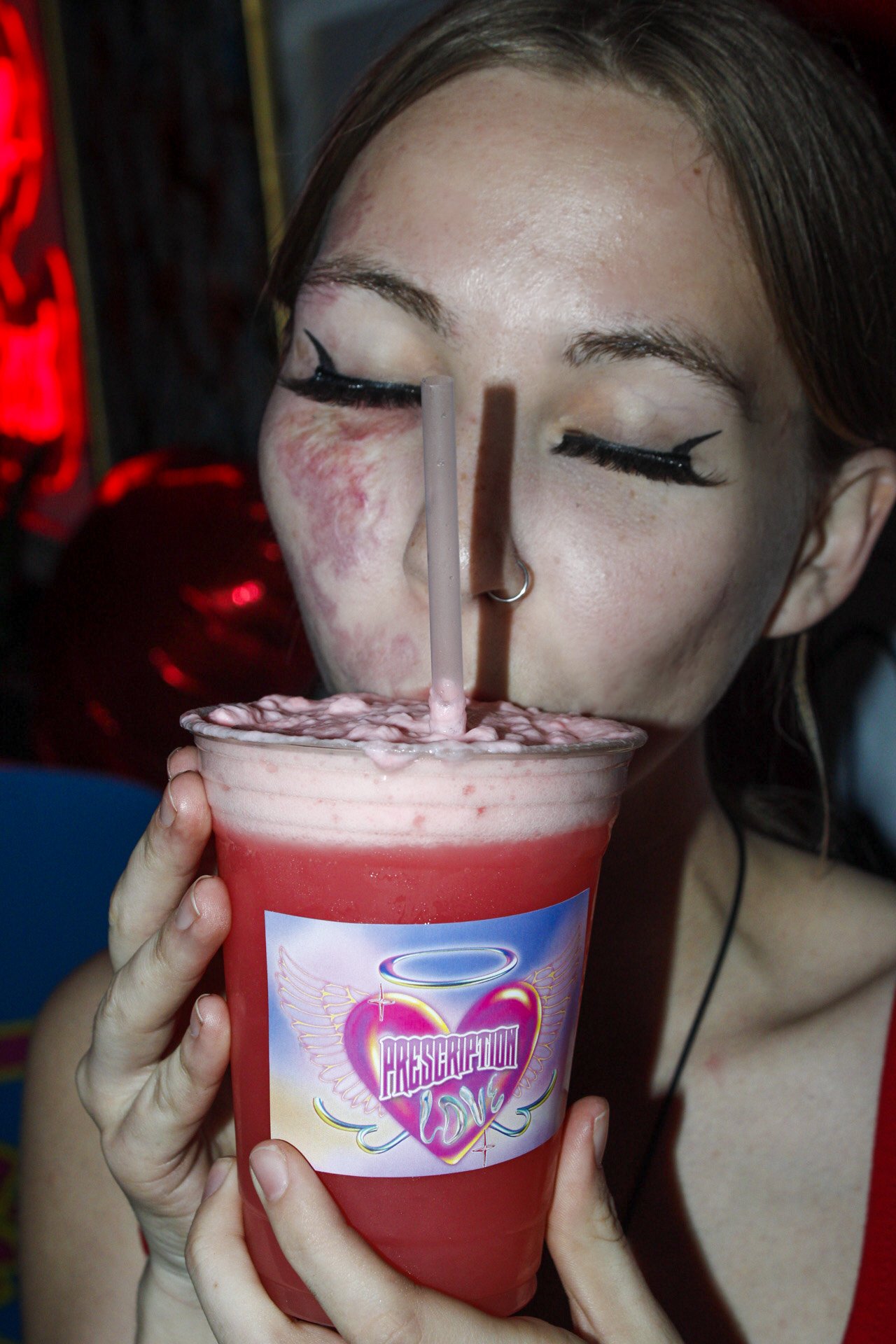 A woman with makeup and a nose ring drinks a pink slushie through a straw, holding a large cup with a 'Passeration Diva' sticker on it.