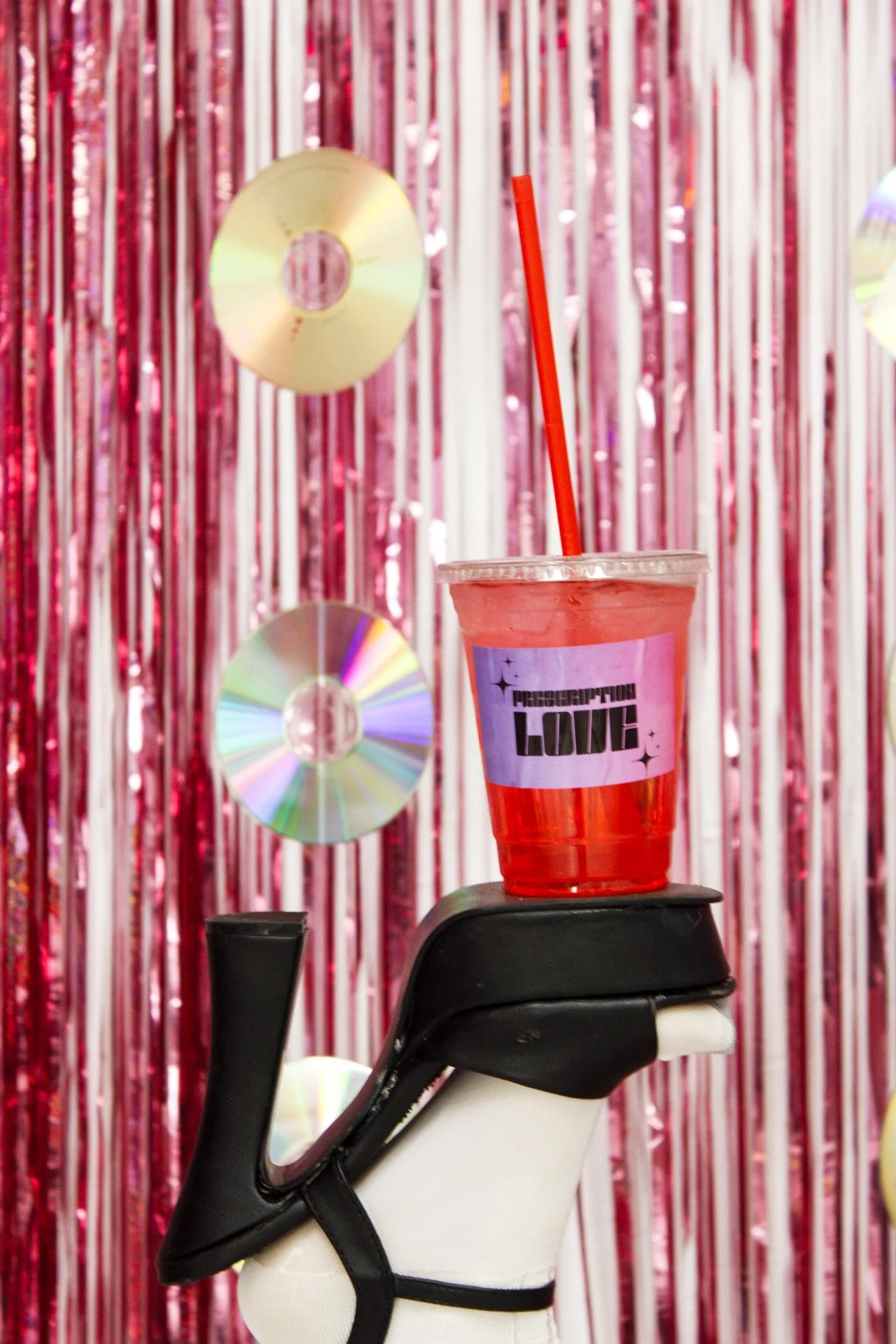 A black and white high-heeled shoe with a red and transparent plastic cup on top containing a pink liquid and a red straw. The cup has a blue label with the words "PROPERCTION LOVE". The background features pink and white metallic streamers and hanging CDs.