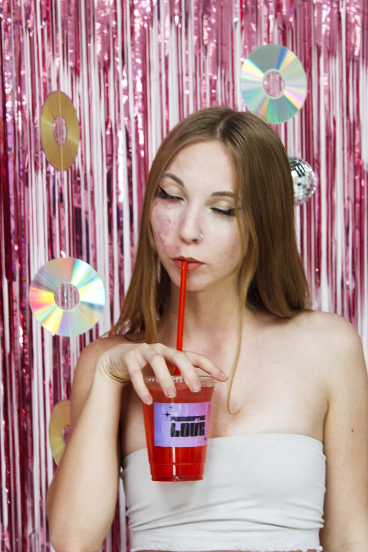 A young woman with long, reddish hair and light skin is drinking a red beverage through a straw. She is standing in front of a pink metallic background with disco ball and CD decorations, wearing a white strapless top.