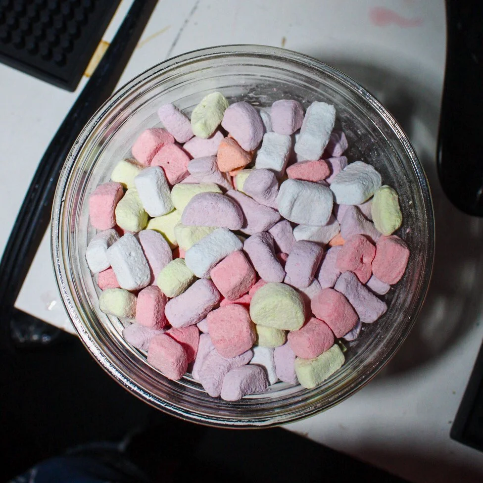 A clear glass jar filled with pastel-colored marshmallows in pink, yellow, purple, and white, placed on a desk or table.
