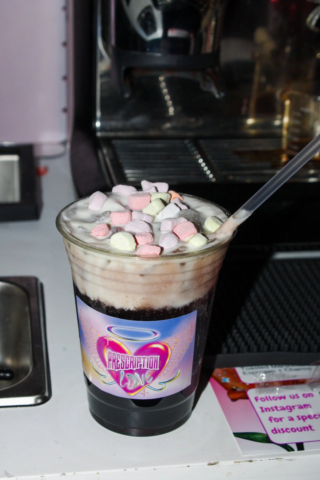 A black soda with whipped cream and mini marshmallows in a clear plastic cup on a counter.