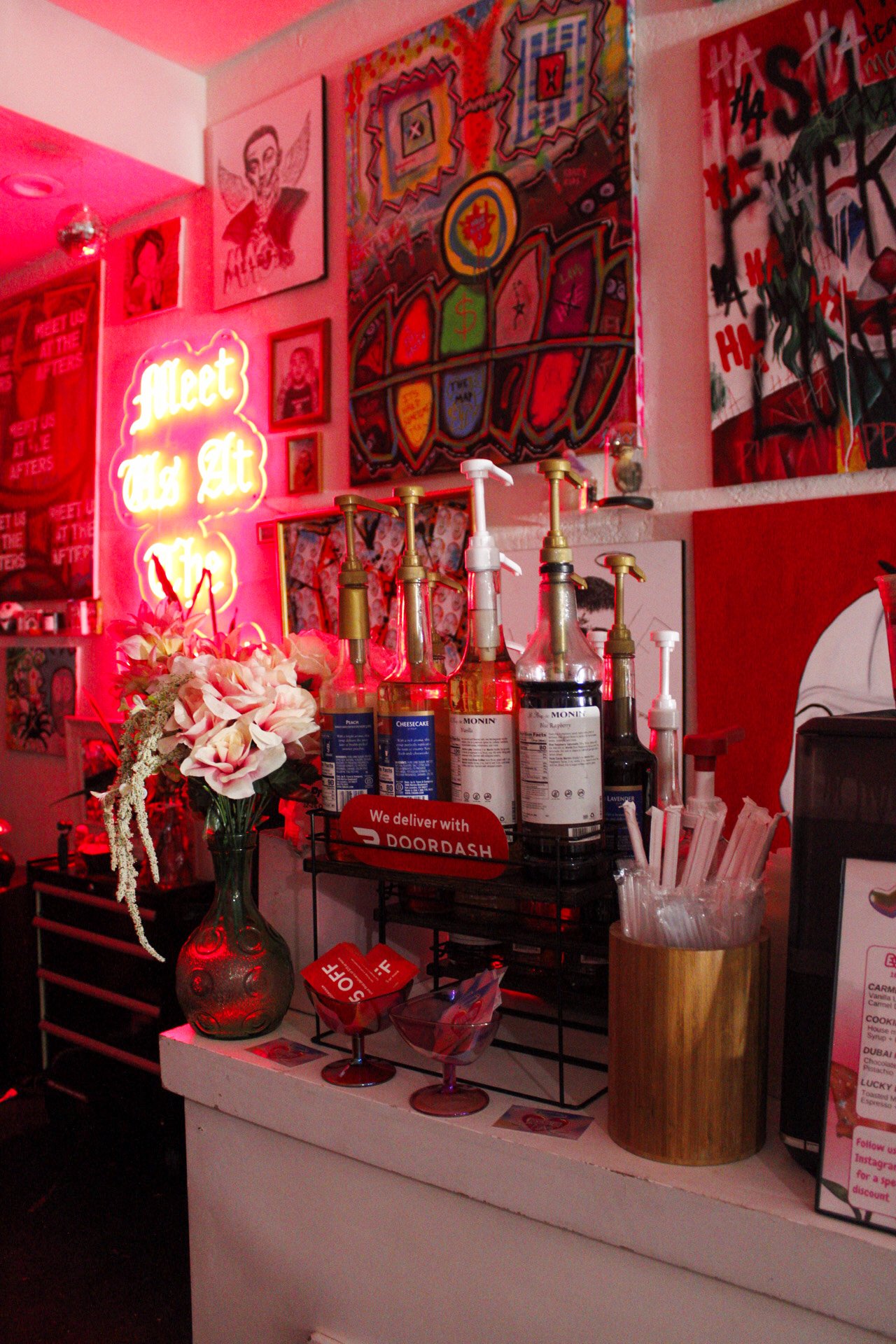 A colorful interior scene featuring multiple abstract and pop art paintings on the wall, a pink neon sign that reads 'Meet Us At The', a vase with pink flowers, various bottles with pump dispensers, a sign indicating delivery service with DoorDash, t