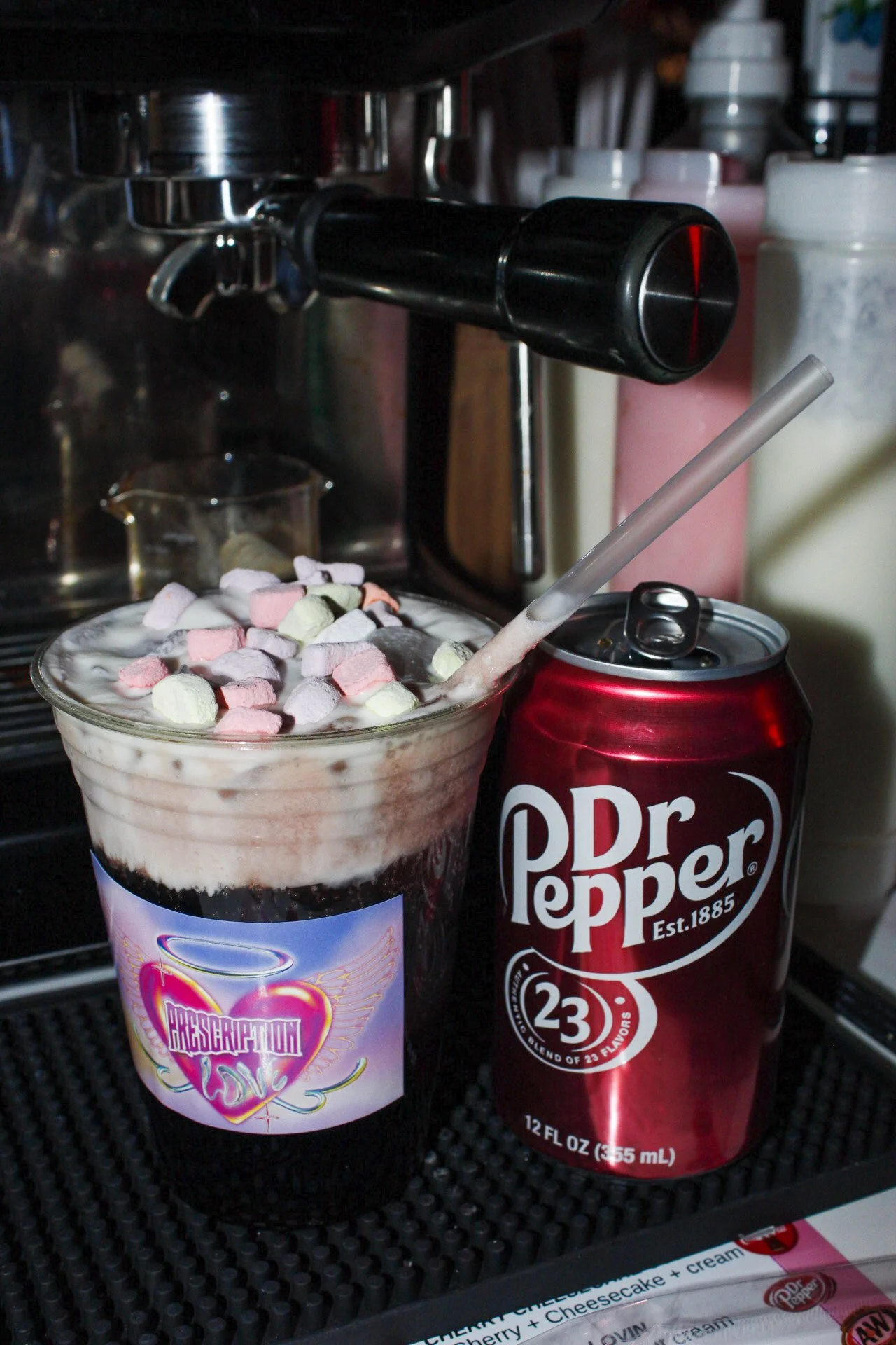 A cup of hot chocolate topped with mini marshmallows, with a straw inserted. Next to it is a can of Dr. Pepper soda.