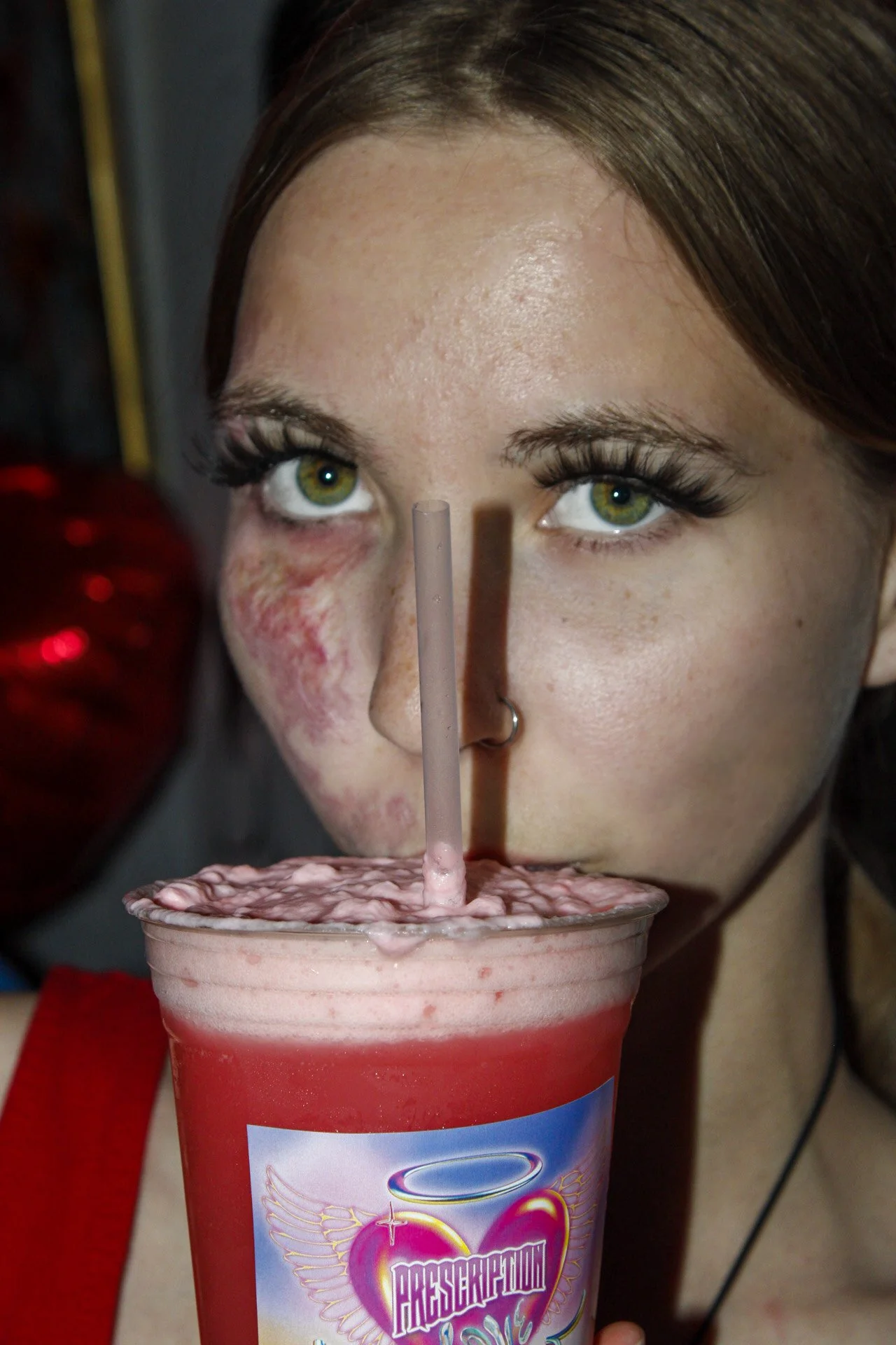 A woman with green eyes and long eyelashes drinks a pink, frothy beverage through a straw, with a logo featuring a heart with angel wings on her cup. She has a serious expression and is wearing a red garment.