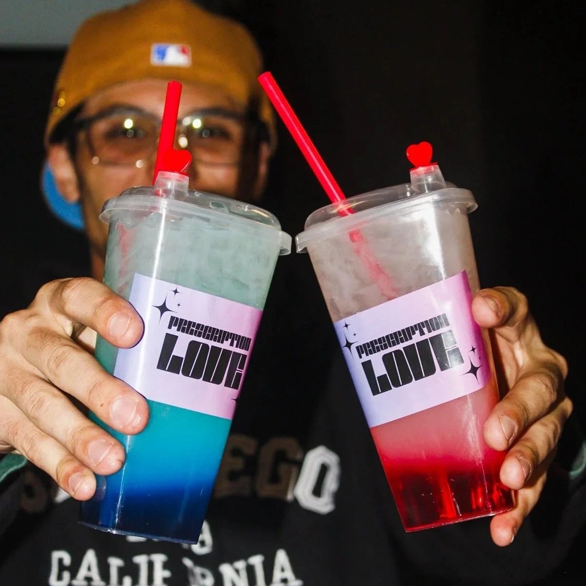 Person wearing glasses and a brown cap holding two colorful drinks with straws, each labeled "PRESCRIPTION LOVE" with a star design.