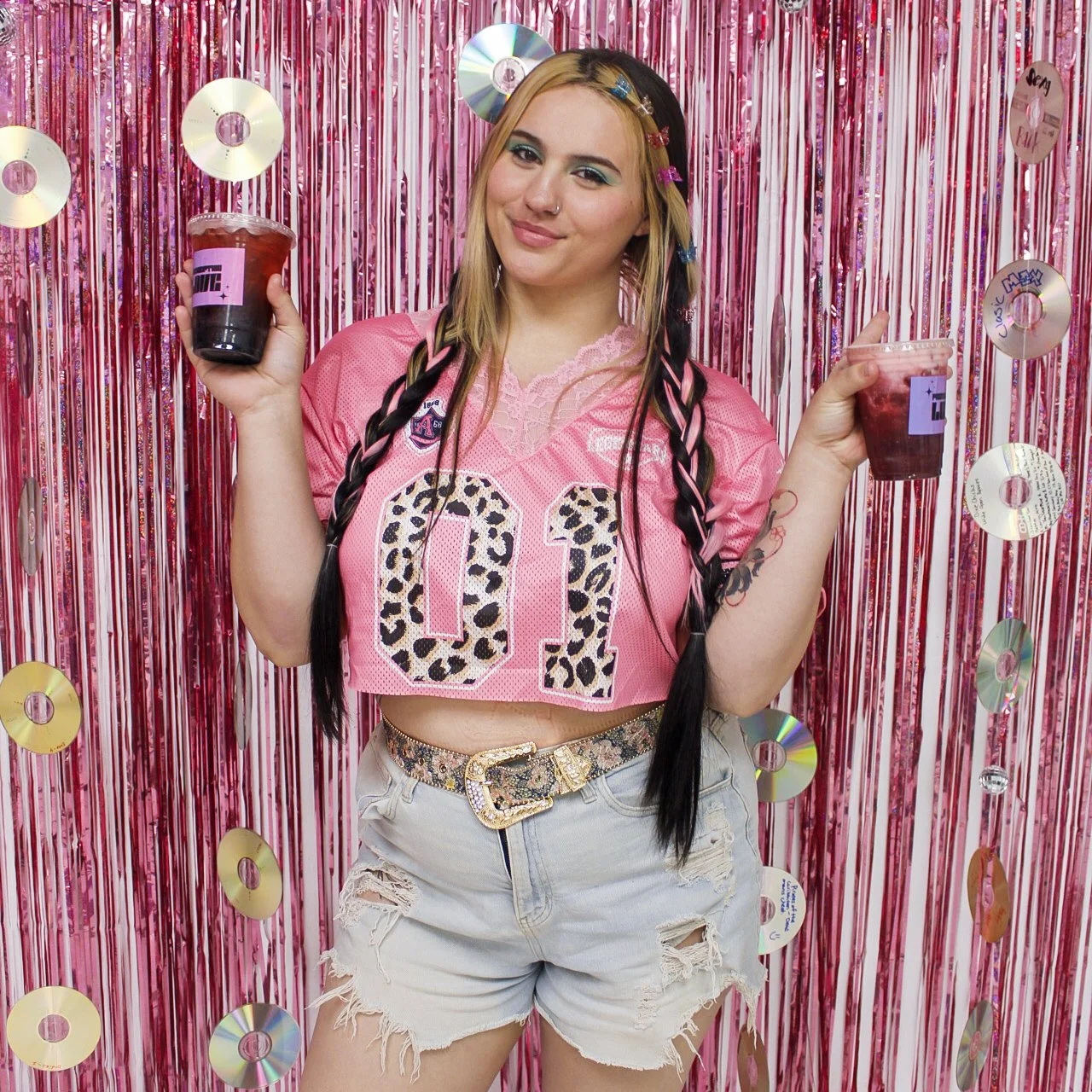 A young woman with long dark hair and light hair, wearing a pink cropped football jersey with the number 01, distressed denim shorts, and a colorful belt, standing in front of a pink and metallic backdrop with rainbow CDs, holding two drinks.