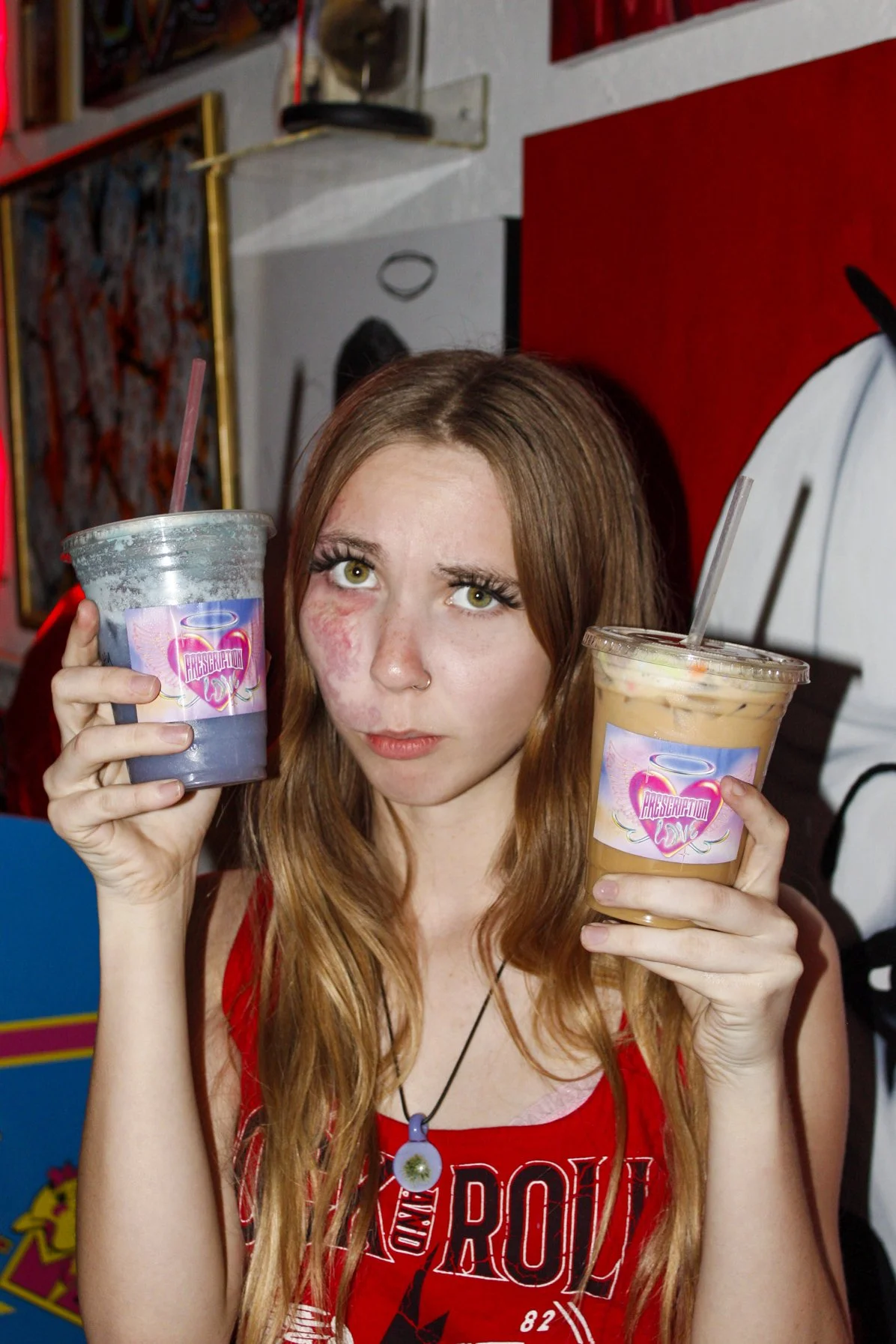 A young woman with long red hair holding two beverages, one purple and one beige, with a serious expression, standing in front of colorful background art.