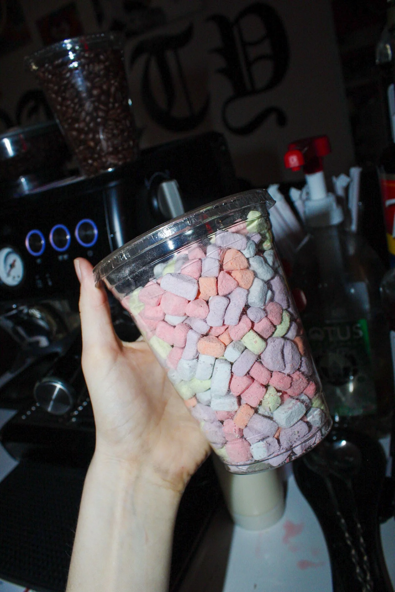 A person's hand holding a clear plastic cup filled with colorful marshmallows, with a coffee maker and various kitchen items in the background.