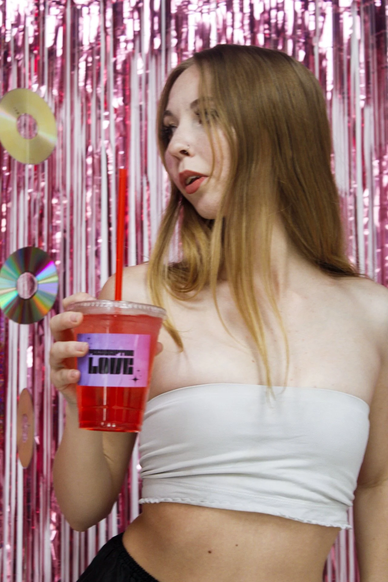 A young woman with red hair holding a red drink with a straw, standing in front of a pink metallic fringe backdrop with CDs hanging on it.
