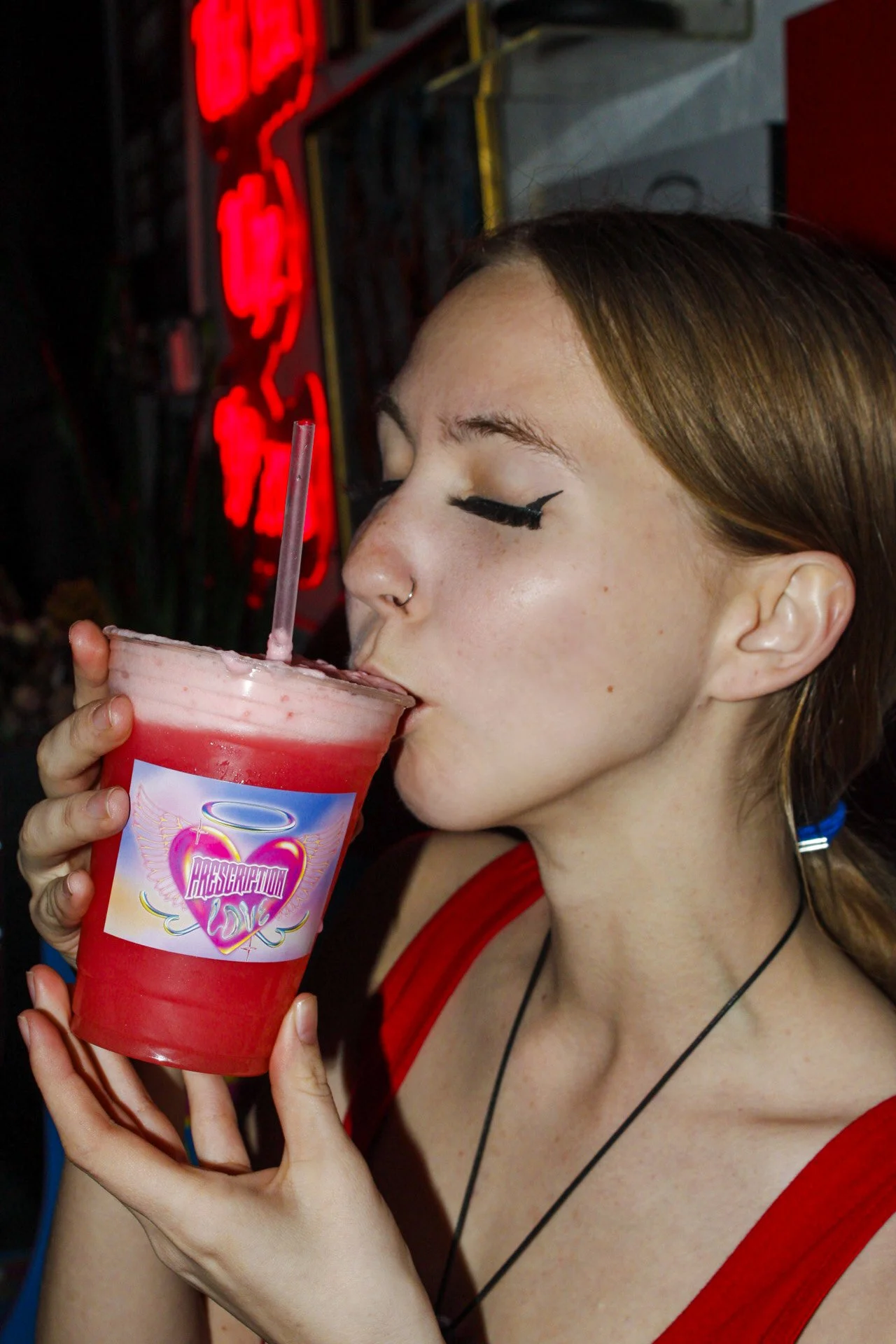 A young woman with light skin, freckles, and brown hair in a ponytail, wearing a red dress and black necklace, drinks a pink smoothie through a striped straw at night with a neon sign in the background.