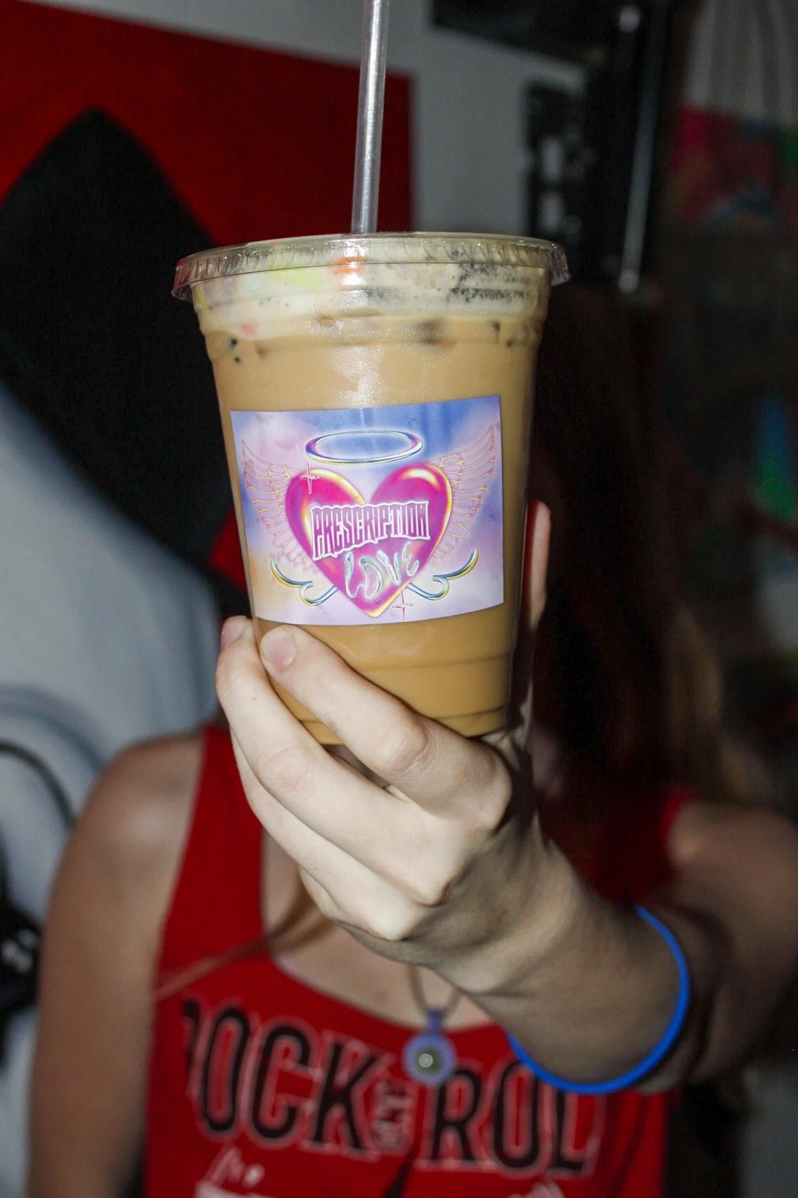 A person is holding a large plastic cup of iced coffee with a lid and straw. The cup has a colorful sticker that says 'Resurrection Love' with a heart, wings, and a halo. The person is wearing a red tank top with black and white text, and a blue brac