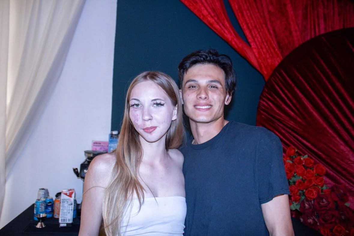 A young man and woman standing close together, smiling at the camera in front of a decorated backdrop with red flowers and fabric.