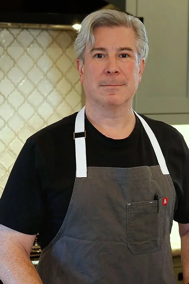 A middle-aged man with gray hair wearing a black t-shirt, a gray apron, and a white apron strap, standing in a kitchen with beige tile backsplash.