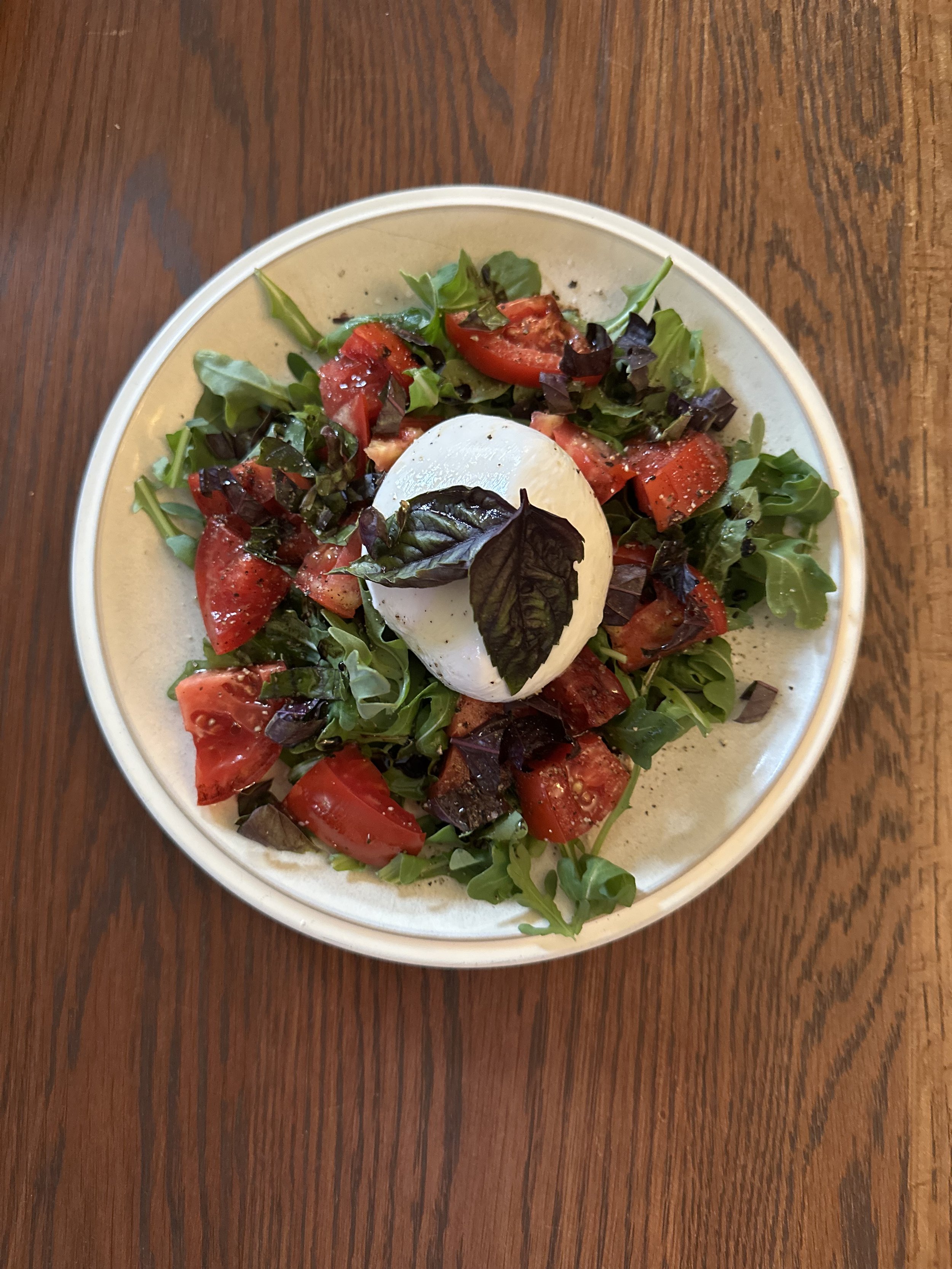 Salad with chopped tomatoes, mixed greens, and a ball of burrata cheese topped with basil leaves on a white plate, placed on a wooden table.