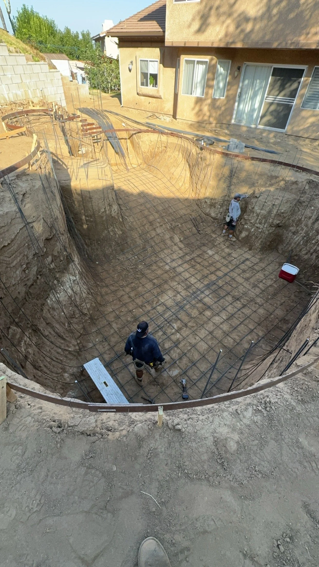 Construction workers are working on the foundation of a building, with rebar grid and excavation site visible in the backyard of a residential house.