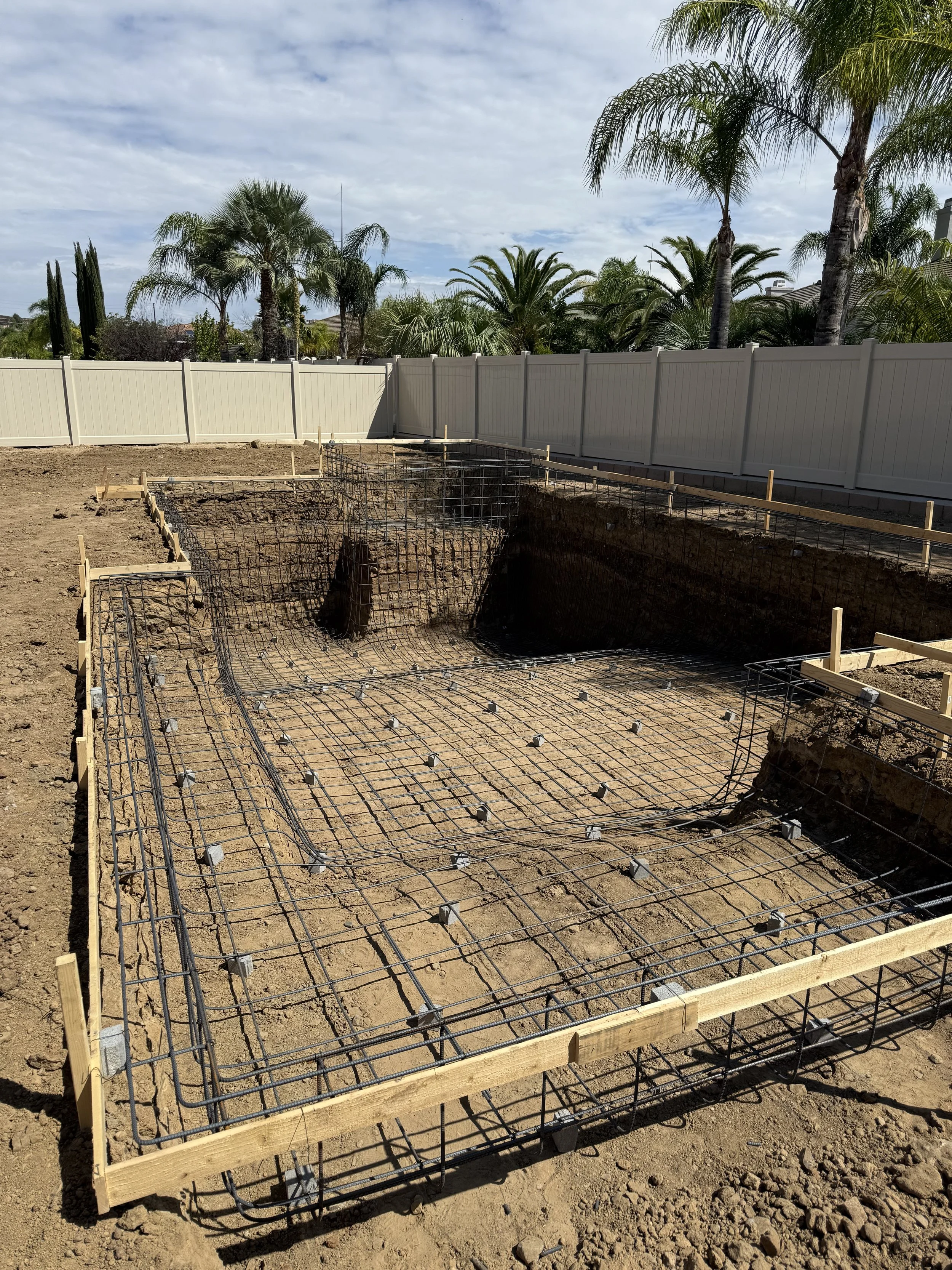 Construction site for a pool with rebar reinforcement and wooden framing, outdoors with trees and a white fence in the background.
