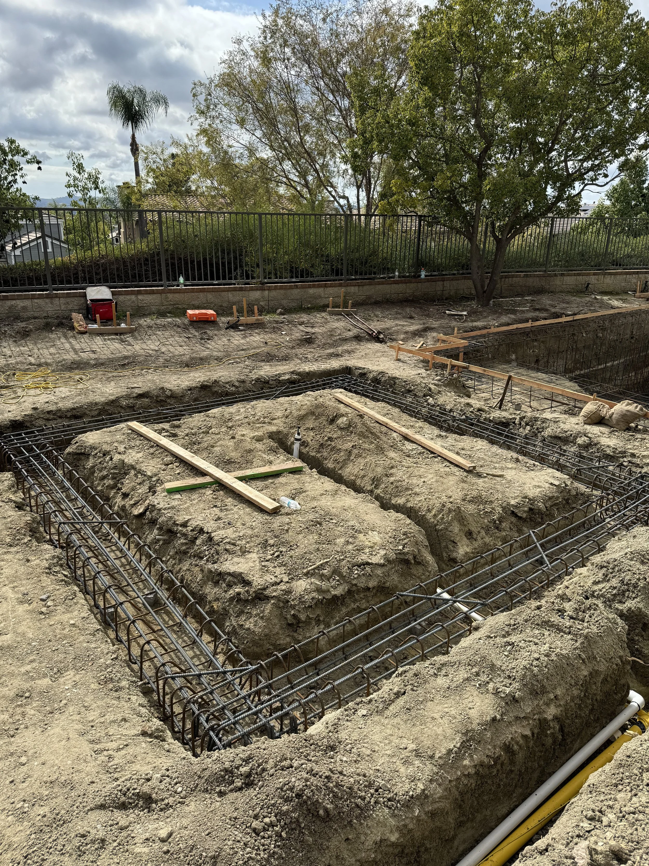 Construction site with dug trenches reinforced with rebar, preparing for foundation pouring, surrounded by dirt and construction tools.