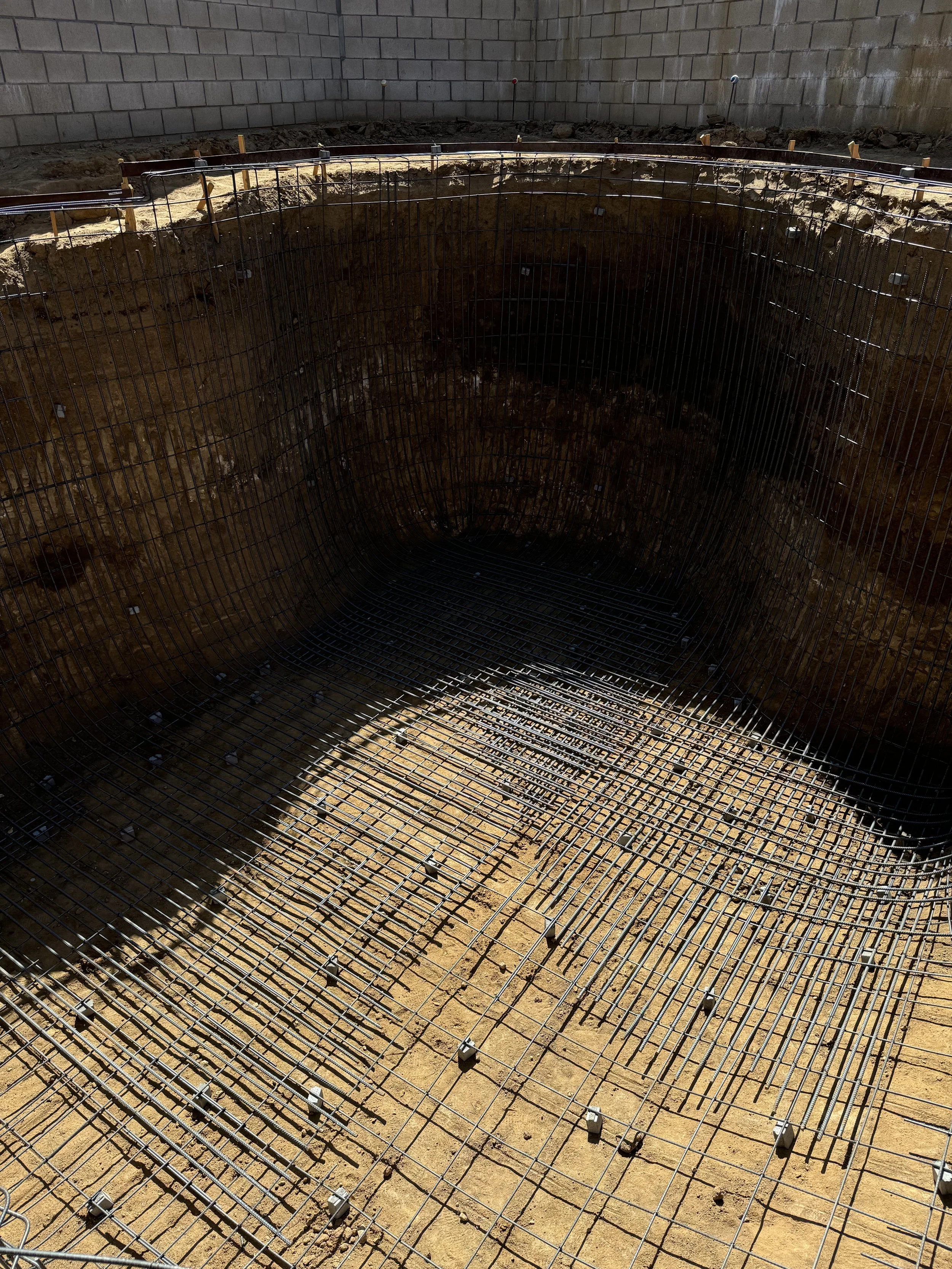 Construction site with a large excavation and steel rebar foundation for a building or structure, surrounded by a concrete wall.