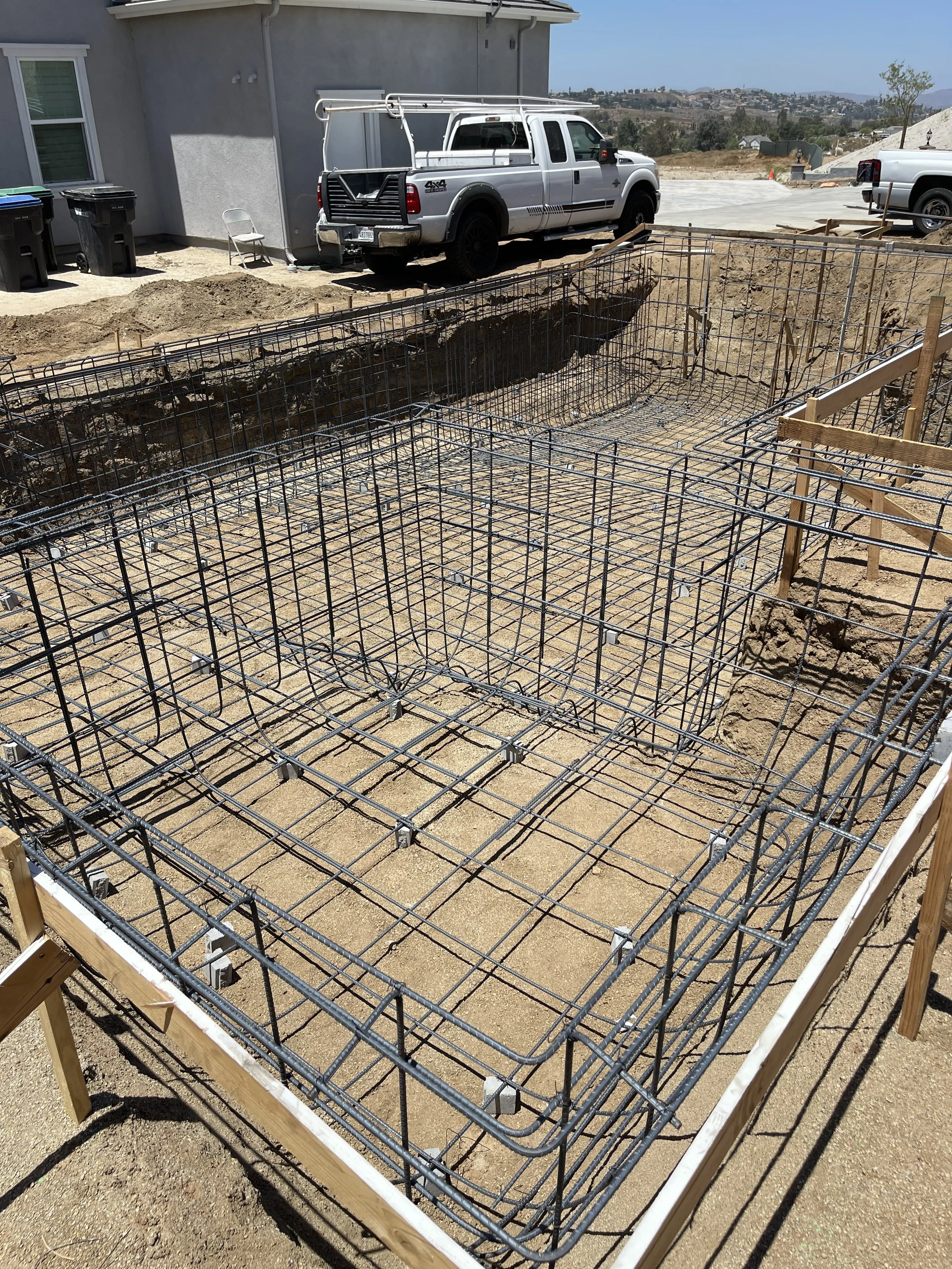 Construction site with metal rebar framework for a foundation, surrounded by wooden stakes, in a residential area under clear skies.