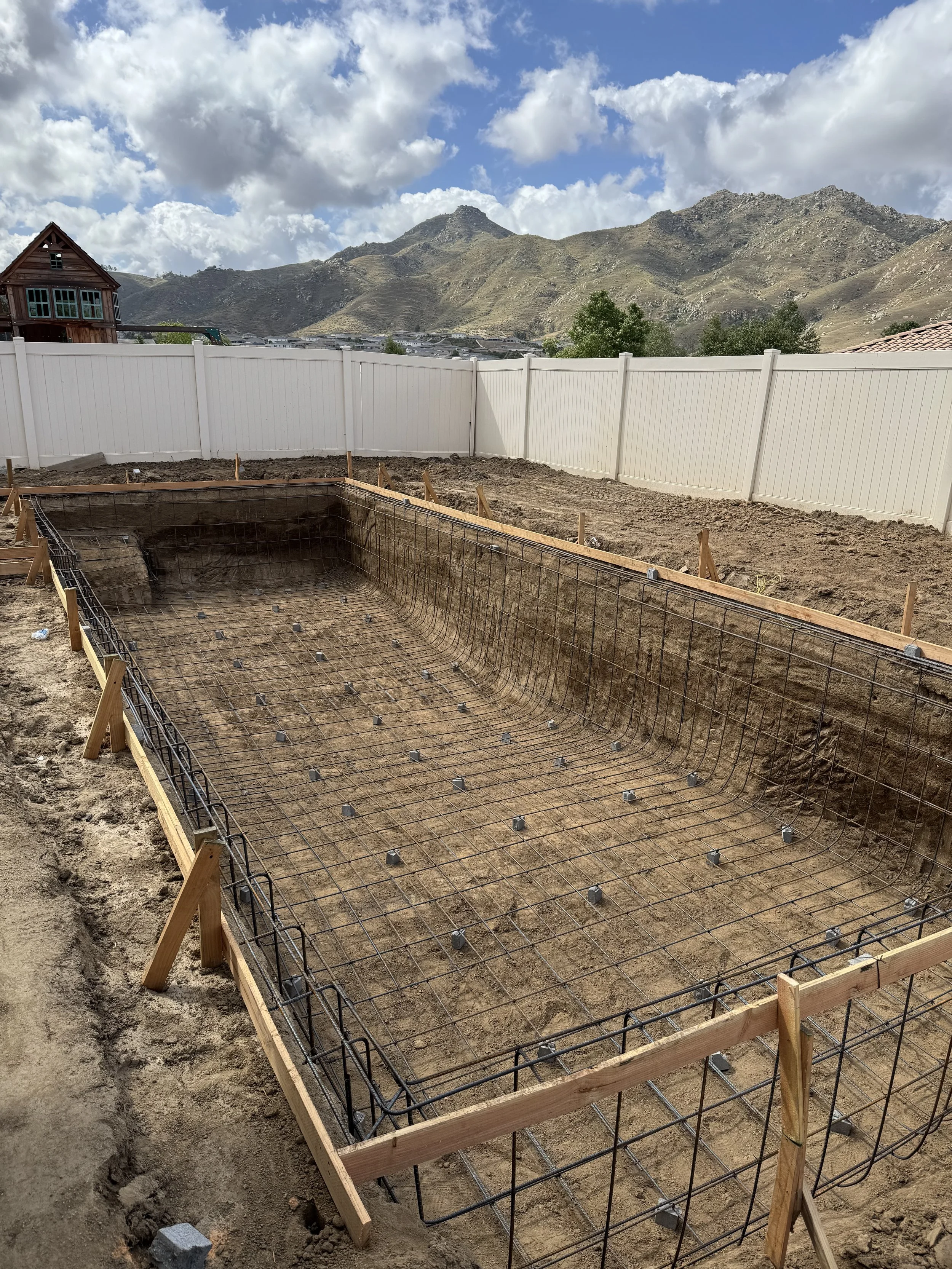 Construction site showing a rectangular foundation with rebar and wooden framing, with a white fence and mountainous landscape in the background.