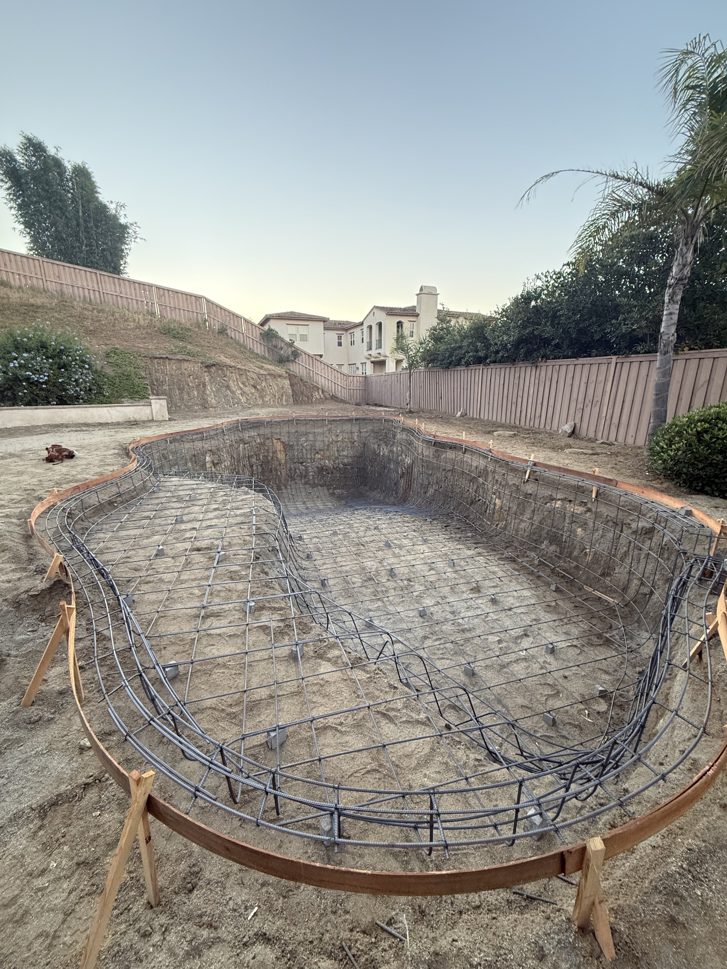 Construction site with an in-ground pool outline and steel rebar reinforcement, surrounded by wooden framing and dirt, in a backyard with trees and residential buildings in the background.