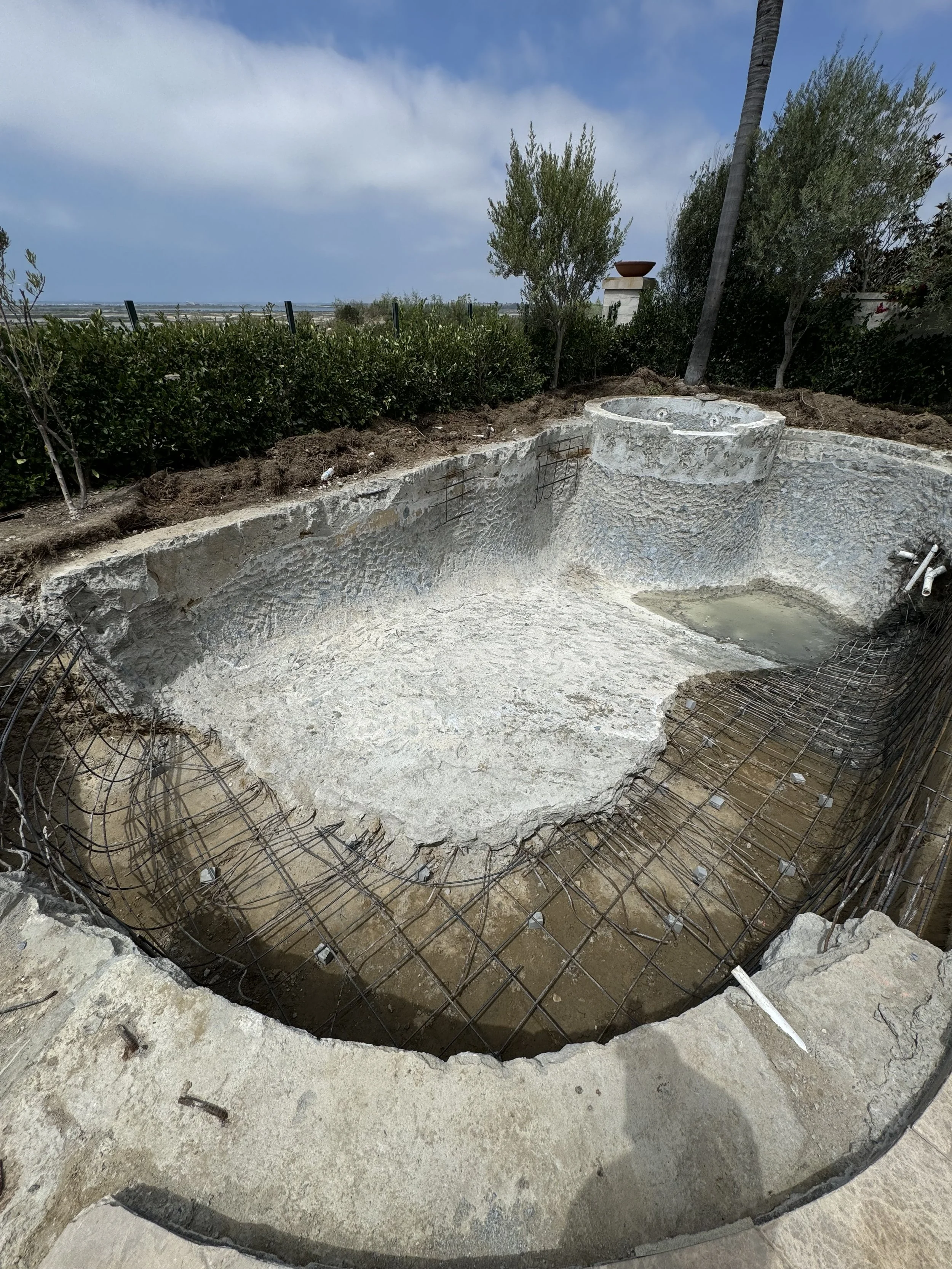 Under construction outdoor hot tub in backyard with rebar reinforcement and plumbing fixtures, surrounded by dirt and bushes.