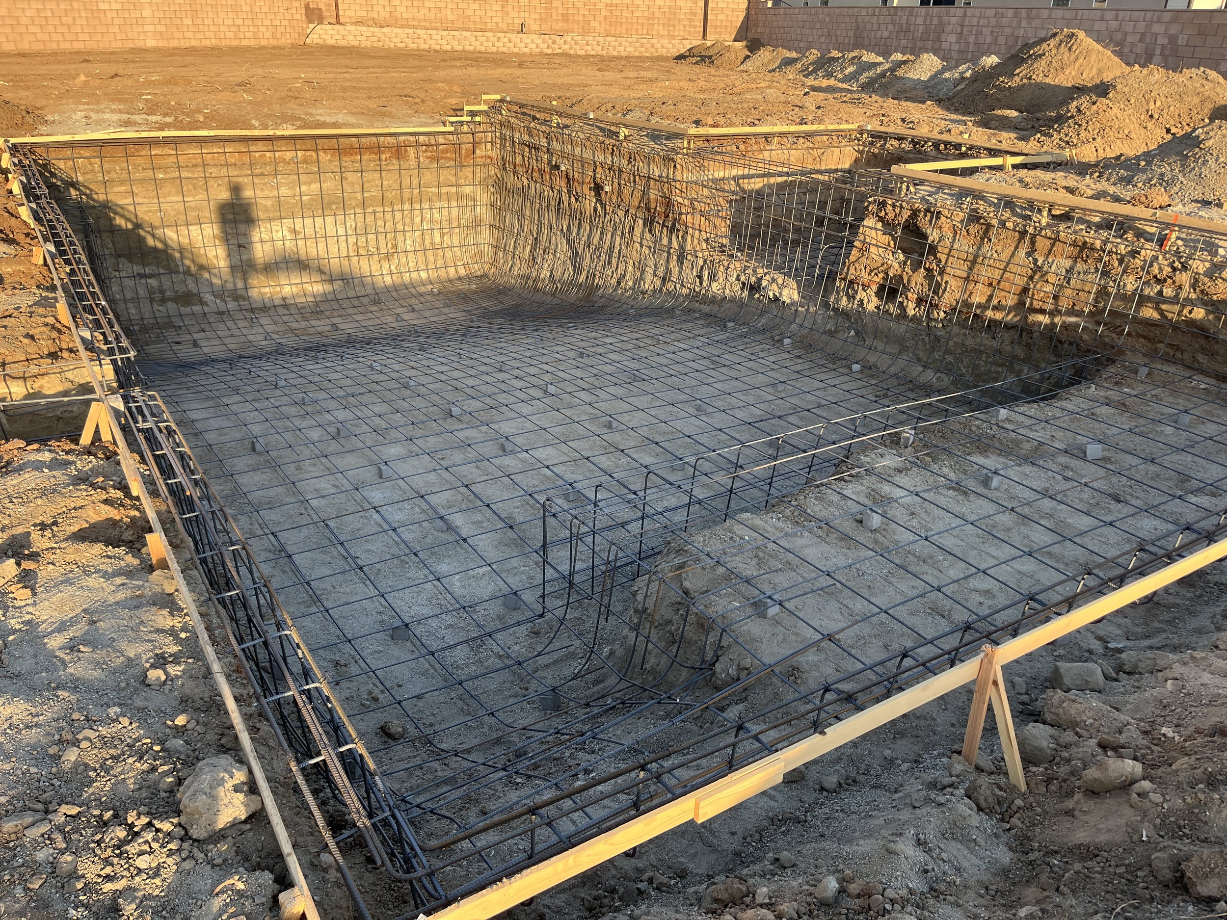 Construction site foundation with rebar grid, wooden framing, and dirt surrounding it at sunset.
