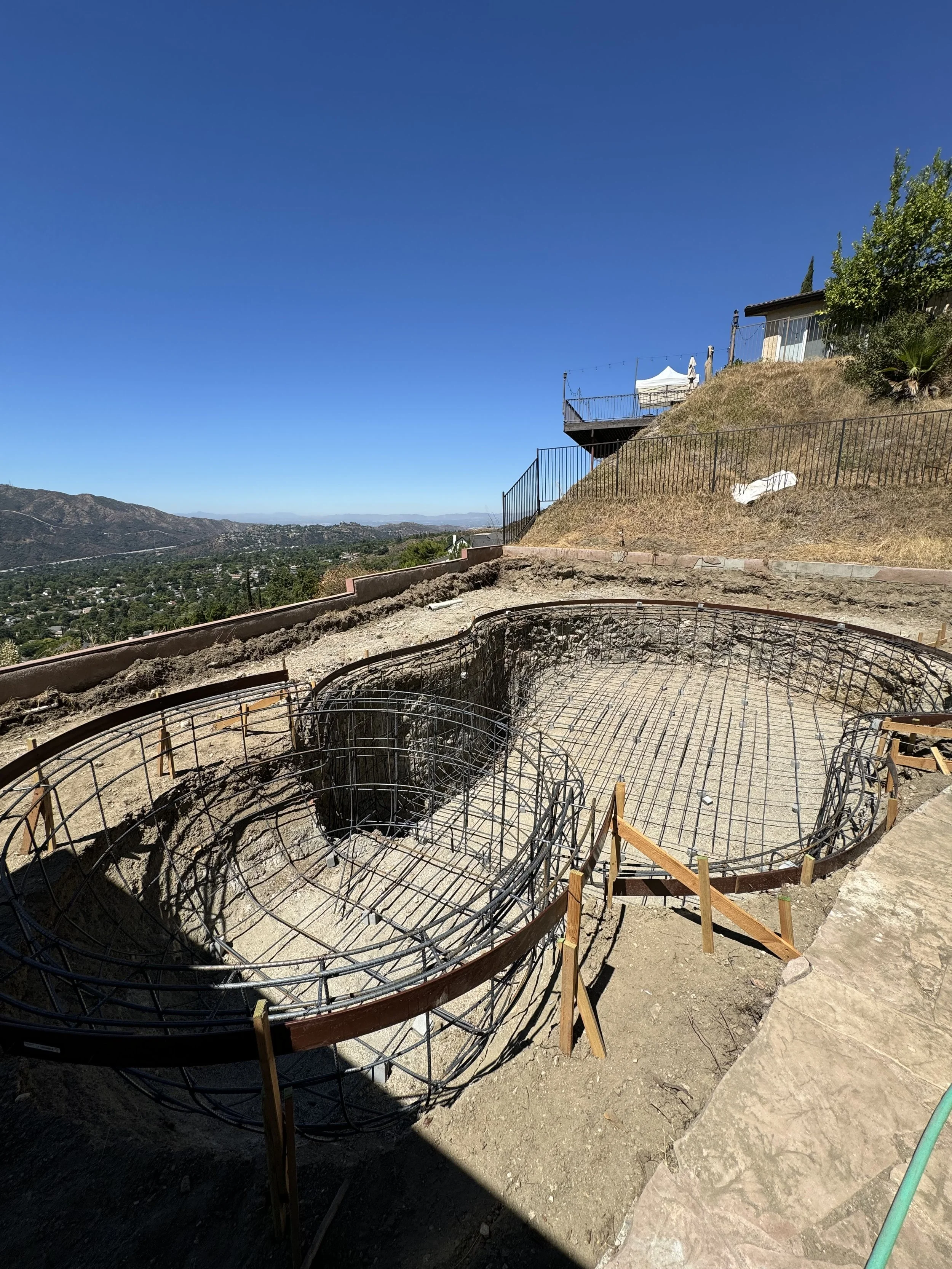 Construction site on a hillside with steel rebar formed into the shape of a swimming pool, with a view of a city and mountains in the distance under a clear blue sky.