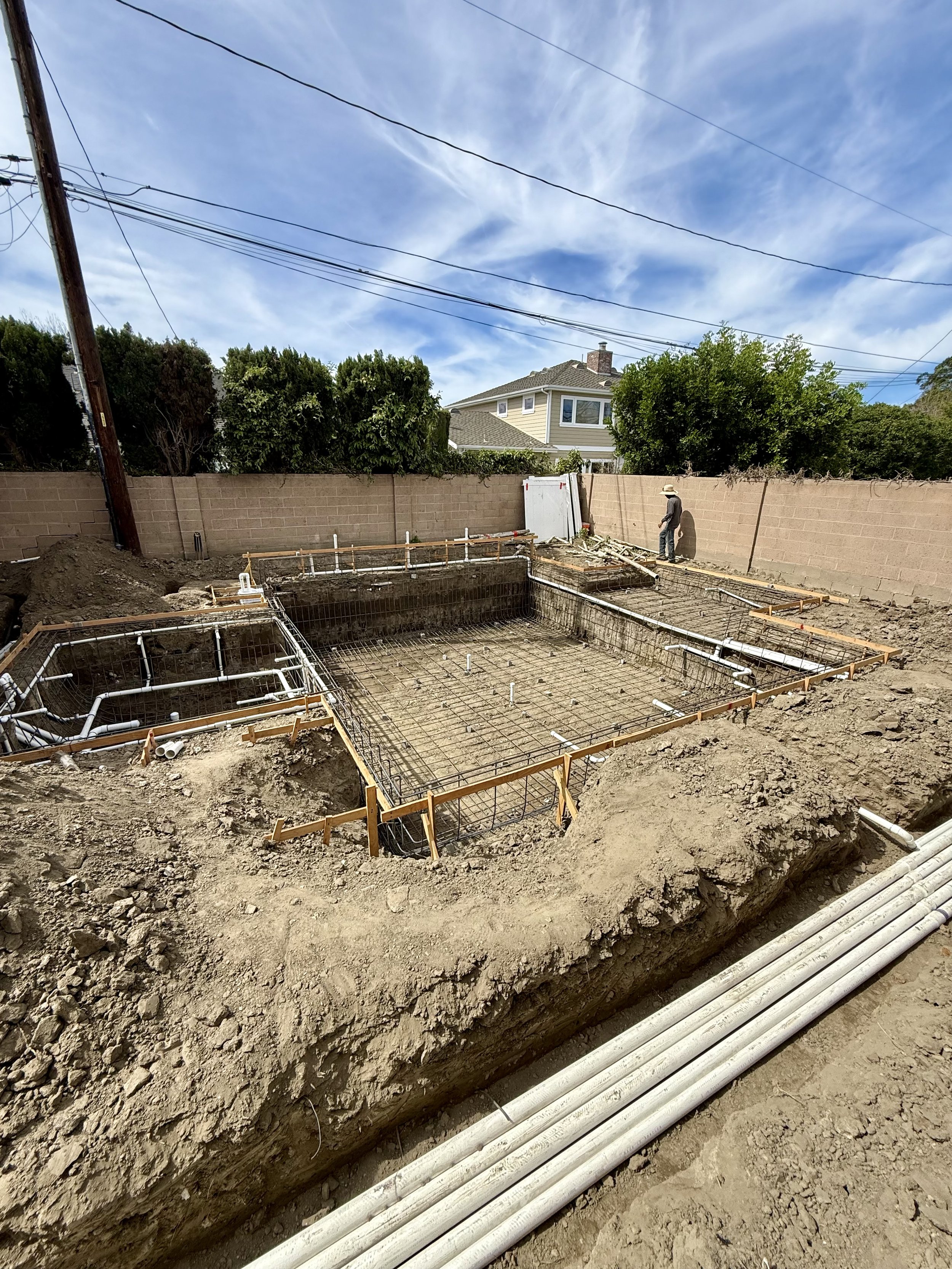 Construction site with a rectangular foundation being built, piping and reinforcement rebar in place, worker standing near a beige wall, a house and trees in the background under partly cloudy skies.