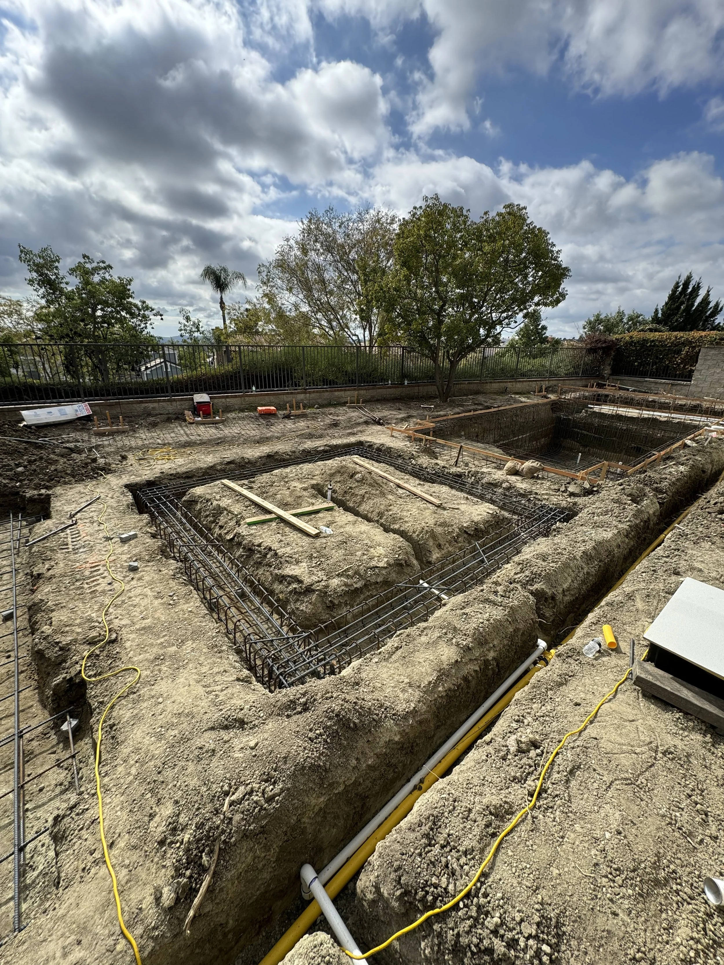 Construction site with foundations being prepared, steel reinforcement bars in excavated trenches, and a partly cloudy sky overhead.